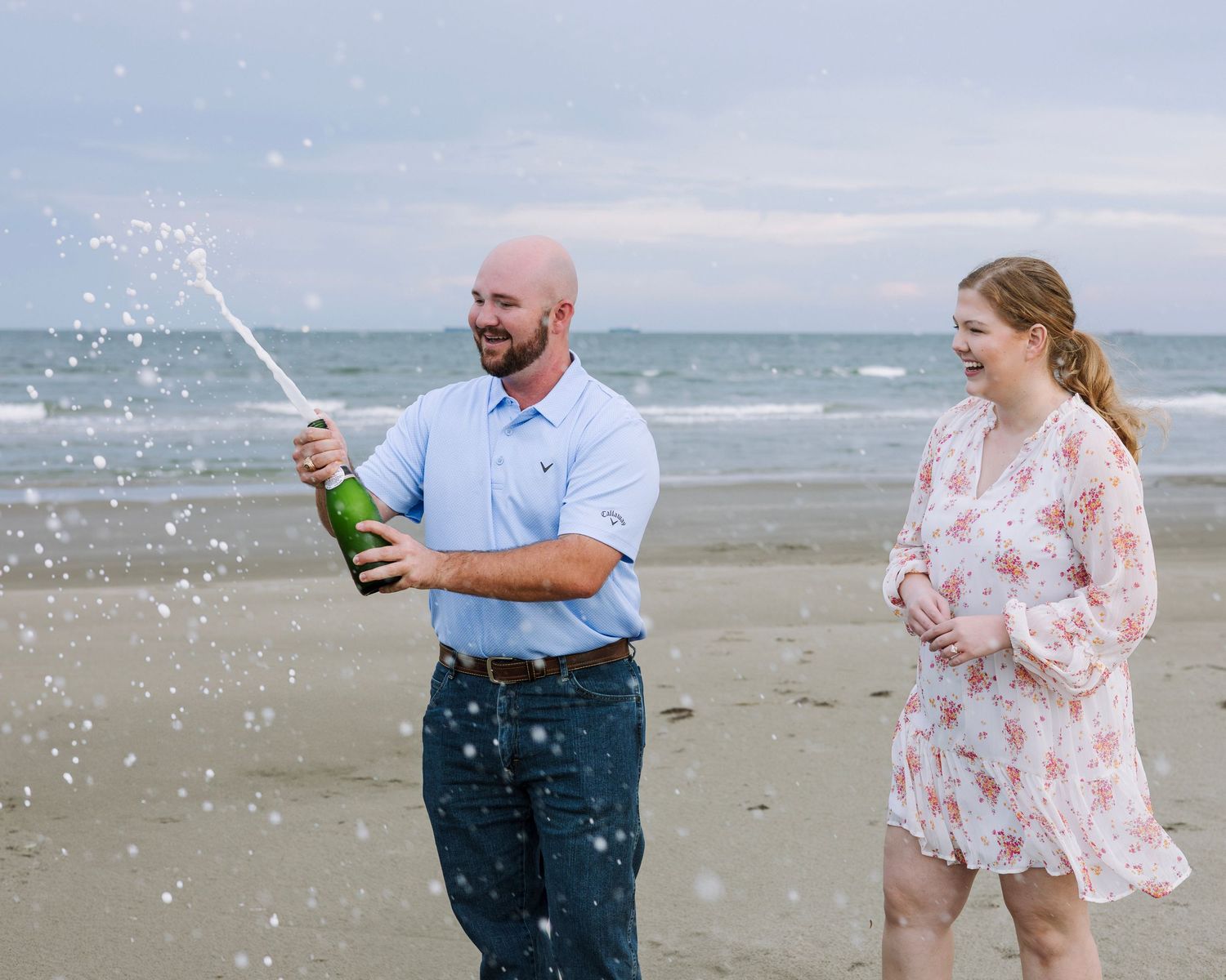 Couple celebrating on beach, man popping champagne bottle, woman in floral dress, ocean waves in background.