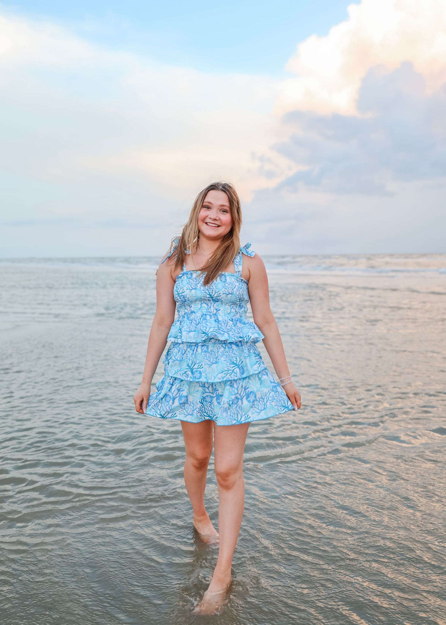 Person wearing light blue ruffled dress wading in ocean water during sunset.