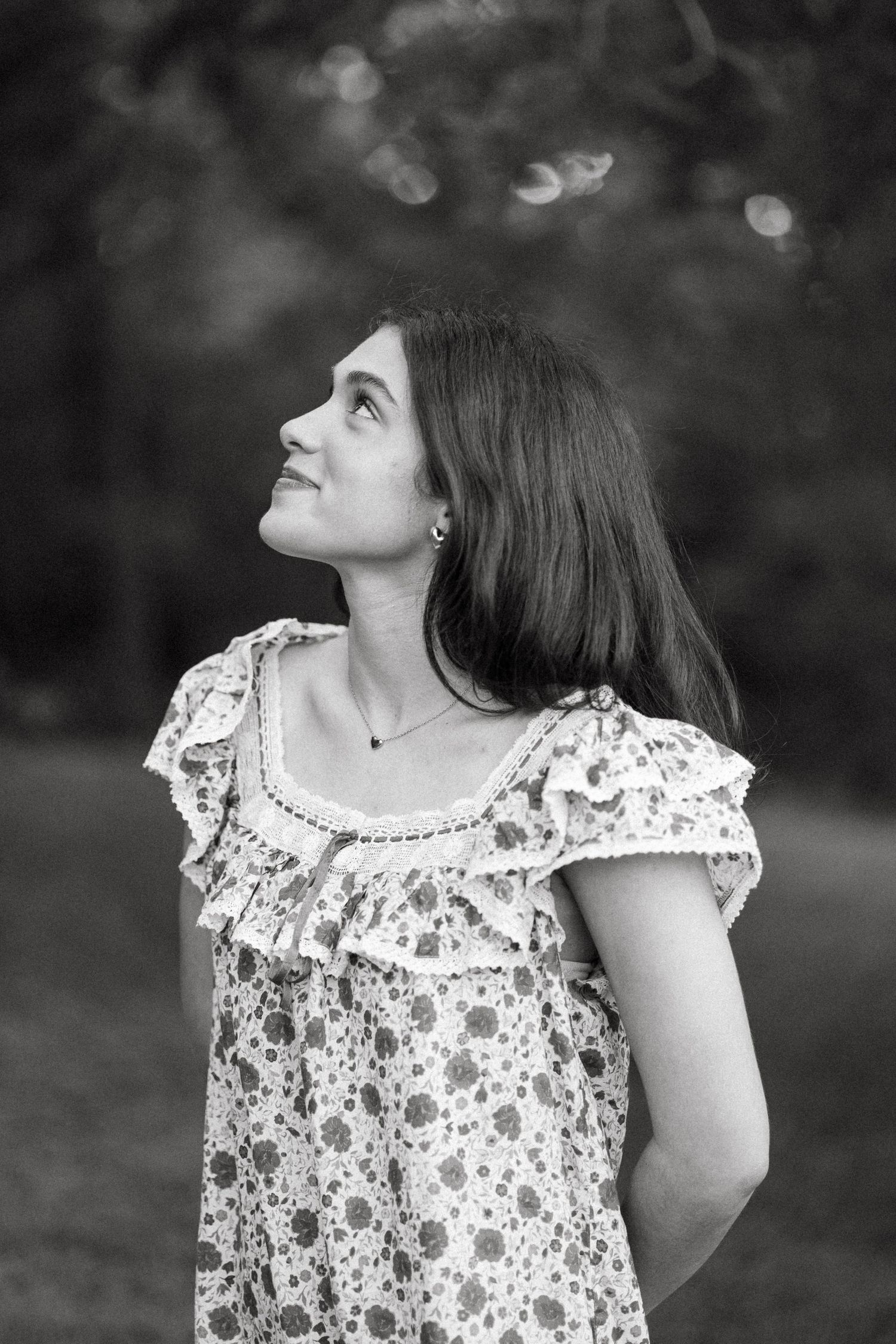 Black and white portrait of a young woman in a floral dress gazing upward outdoors.