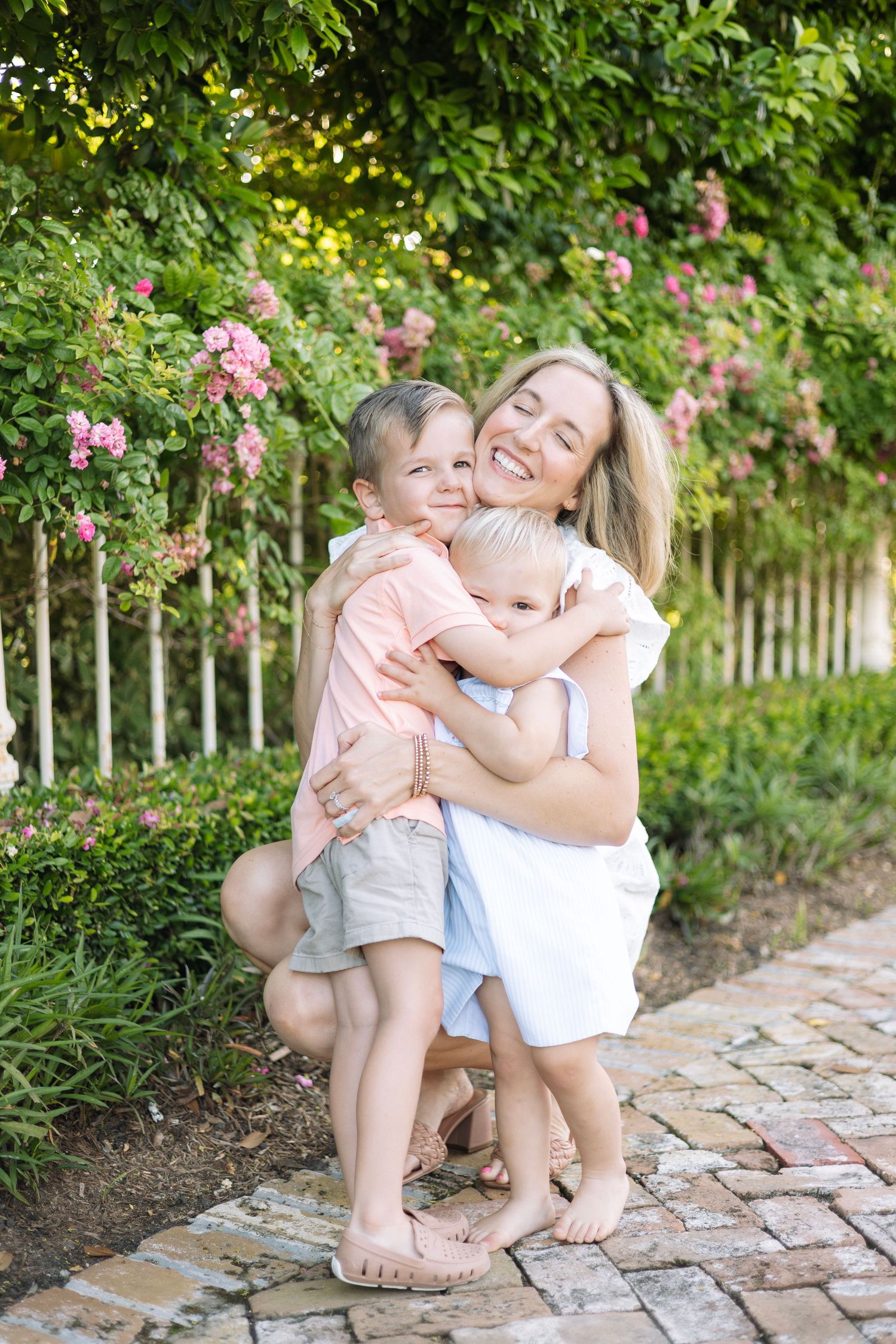 A smiling mother crouches to hug her two young sons in a lush garden with pink roses.