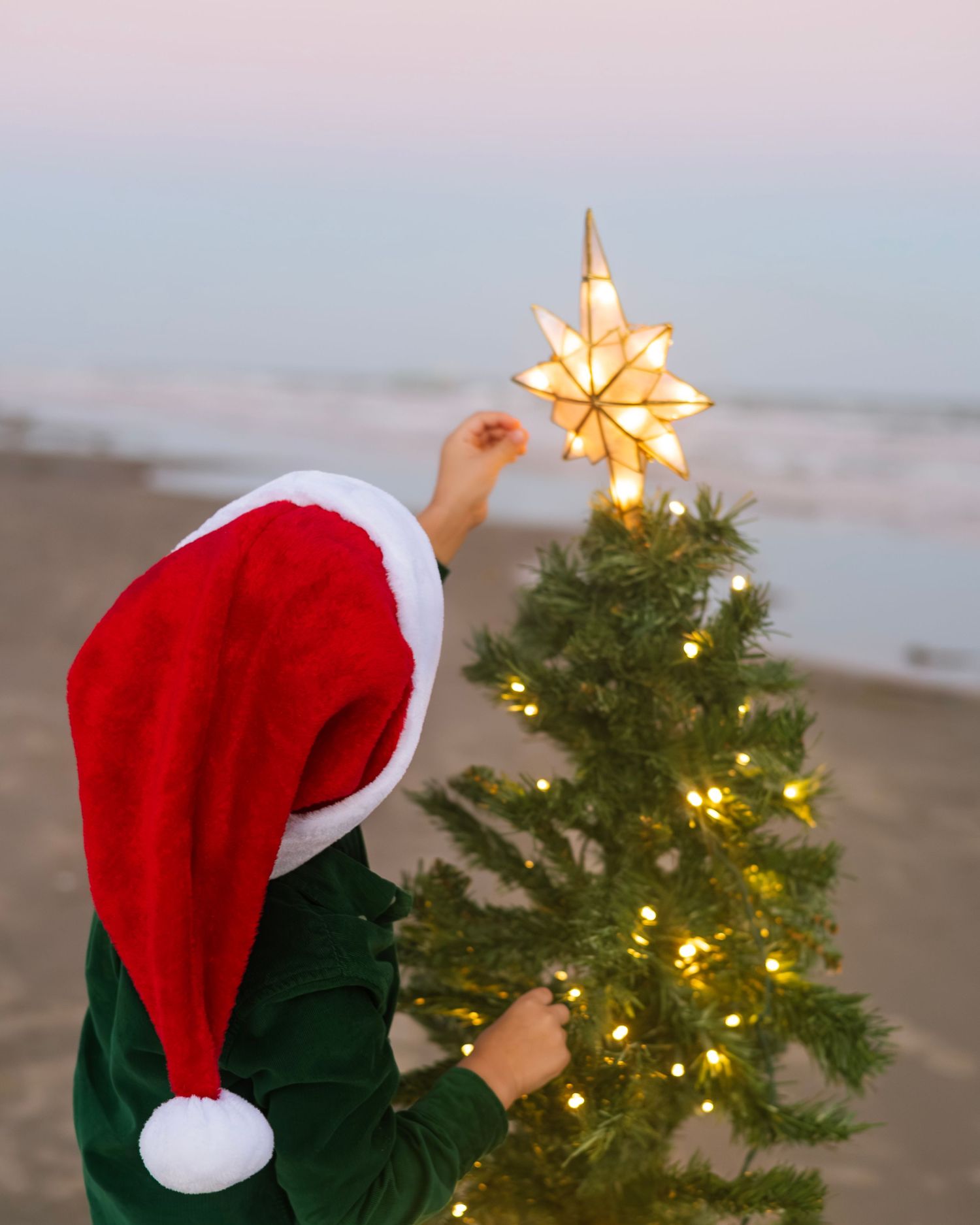 Person in Santa hat reaching up to place star on illuminated Christmas tree at beach during sunset.