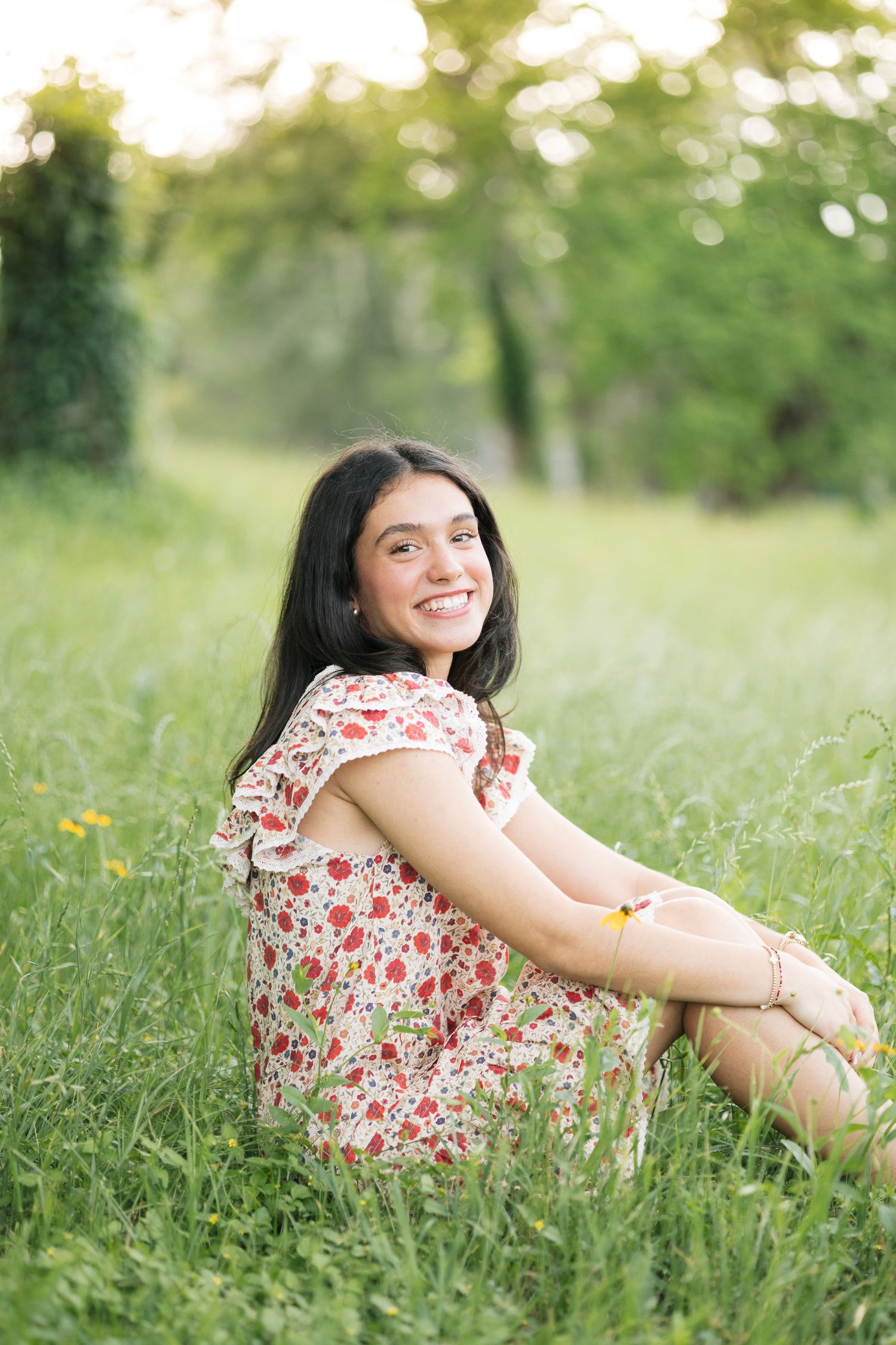 Young woman in floral dress sitting in green grass field smiling outdoors during golden hour.