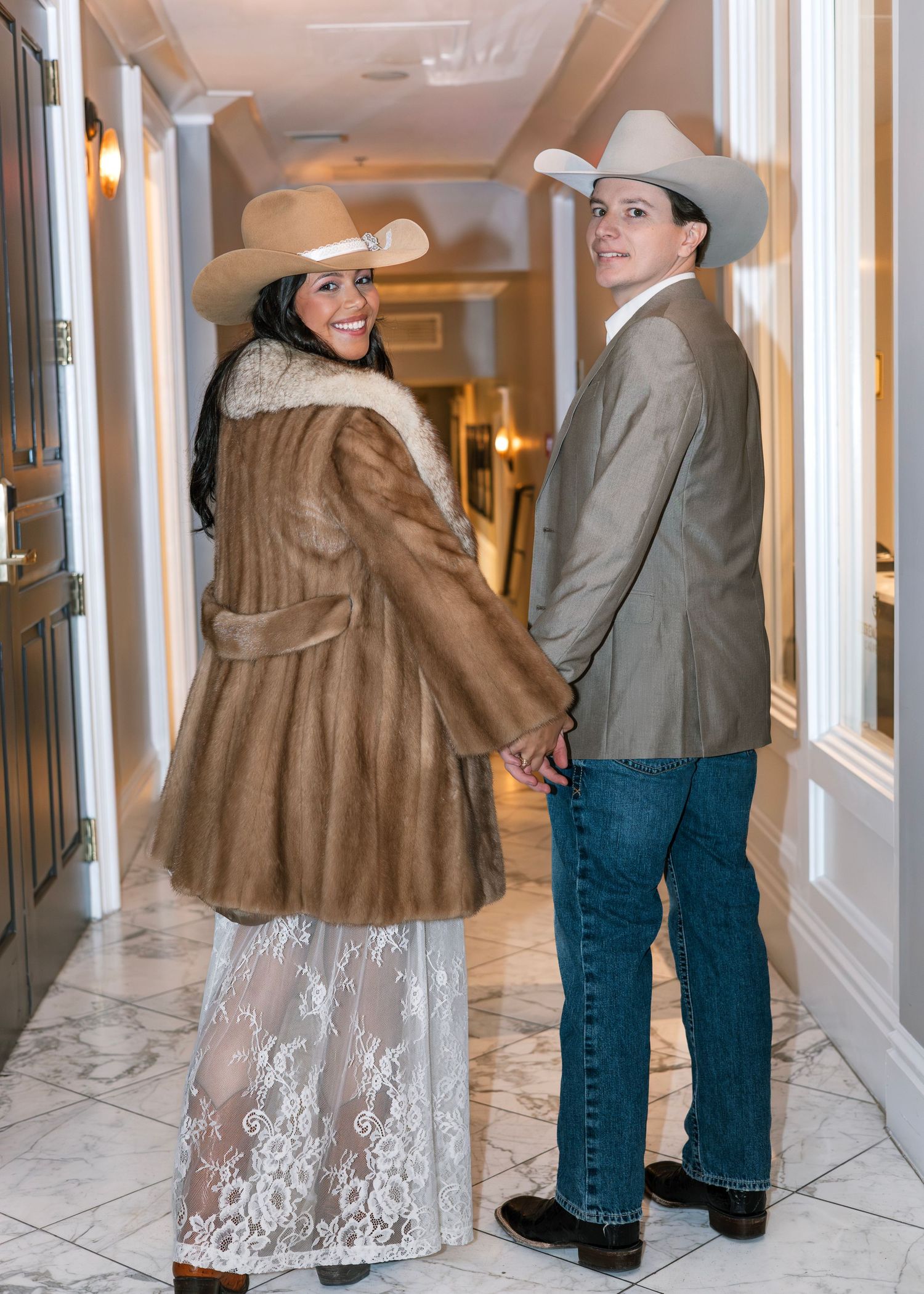 Couple in cowboy hats holding hands in hallway, woman in fur coat and lace skirt, man in blazer and jeans.