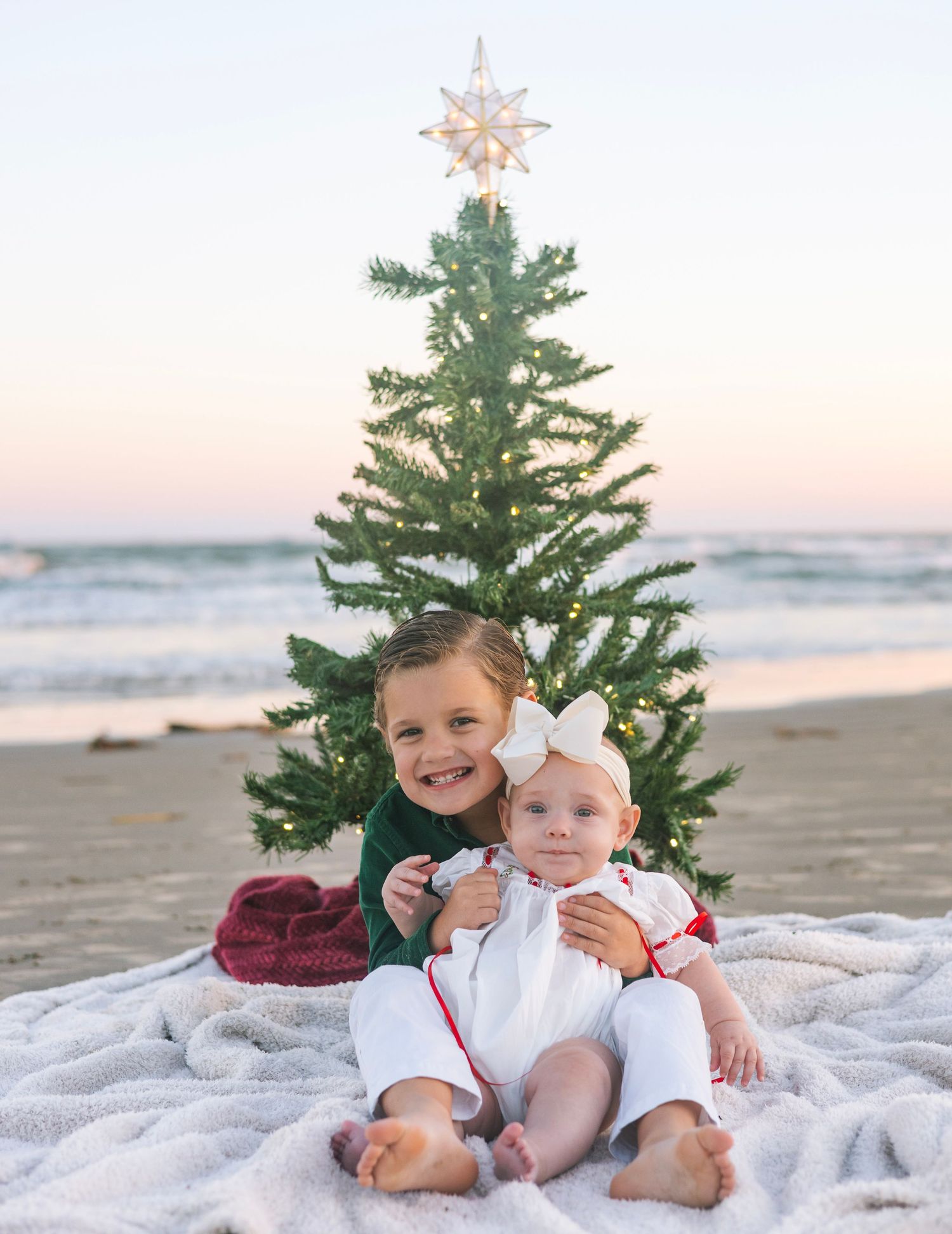 Children laying on a white blanket by a small Christmas tree on the beach at sunset.