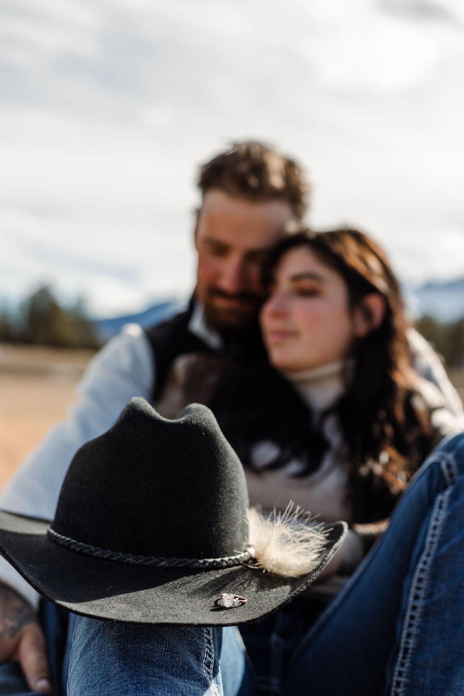 couple poses with cowboy hat in the foreground with the ring on the brim