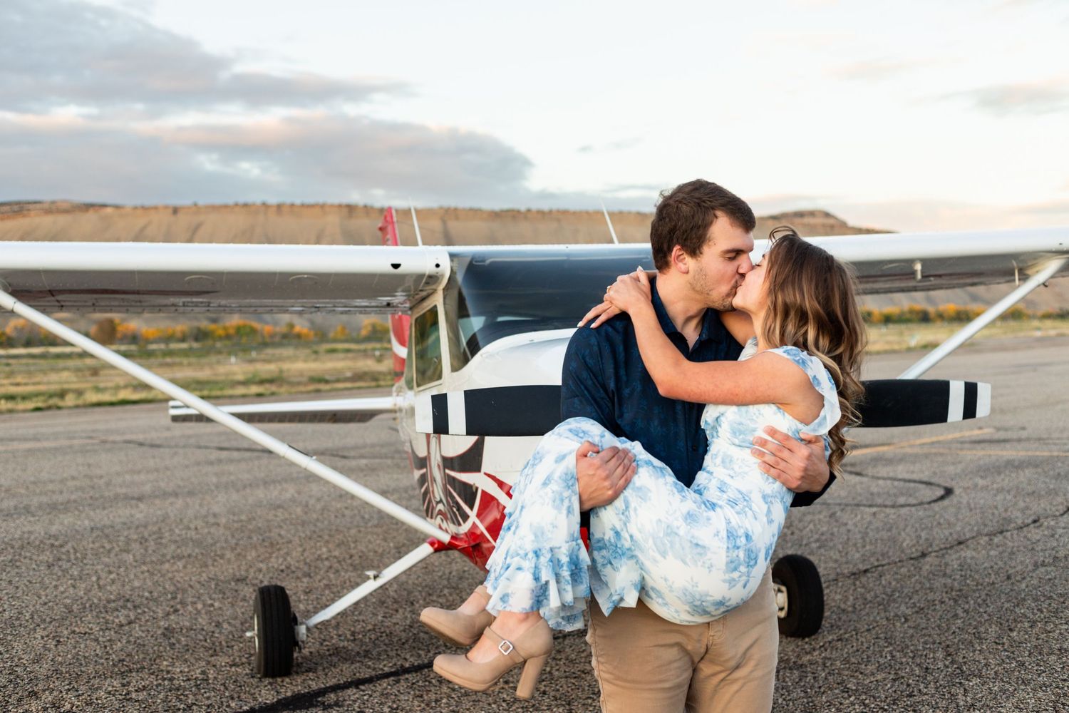 man lifts fiance and kisses her in front of small airplane