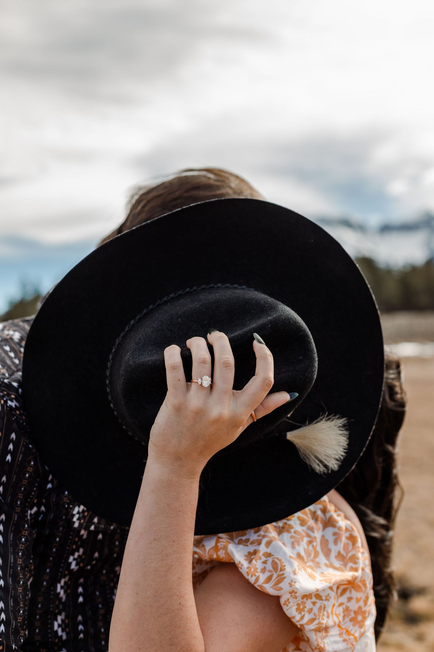 Couple kisses behind cowboy hat but ring is shown with the hand holding the hat