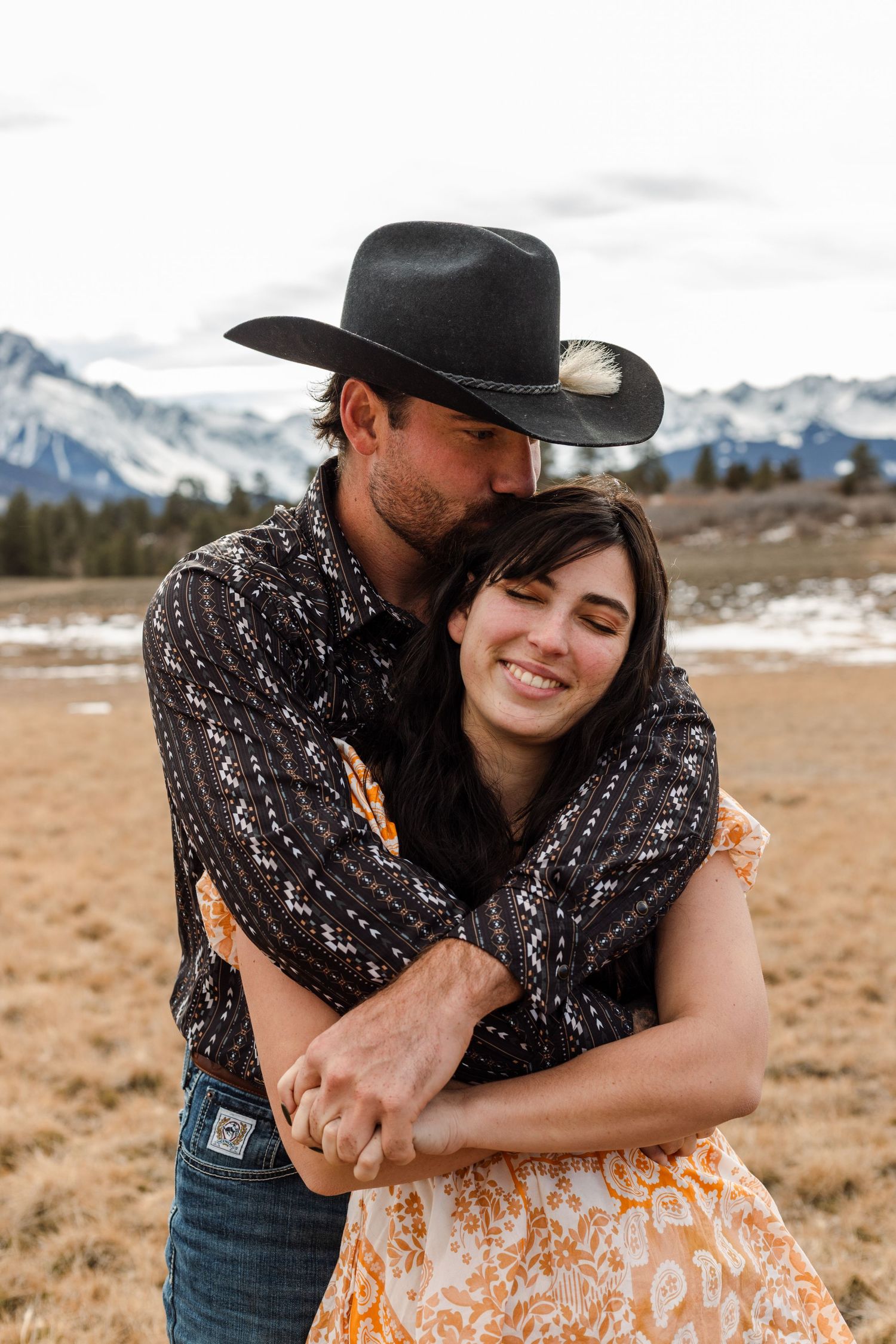 man kisses fiances head in front of mountain background