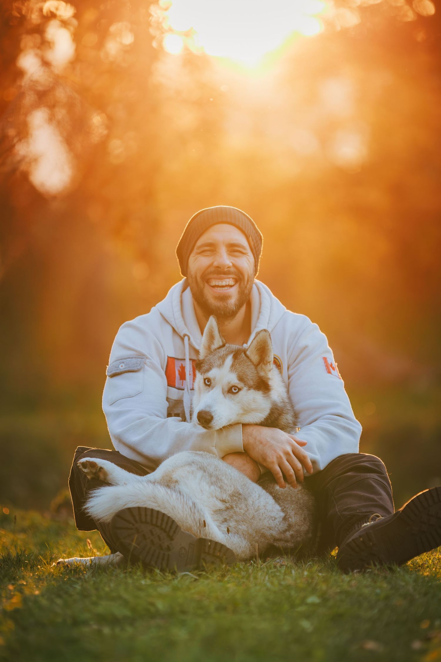 Person and dog share tender moment in warm sunset light while sitting on grass in matching white outfits.