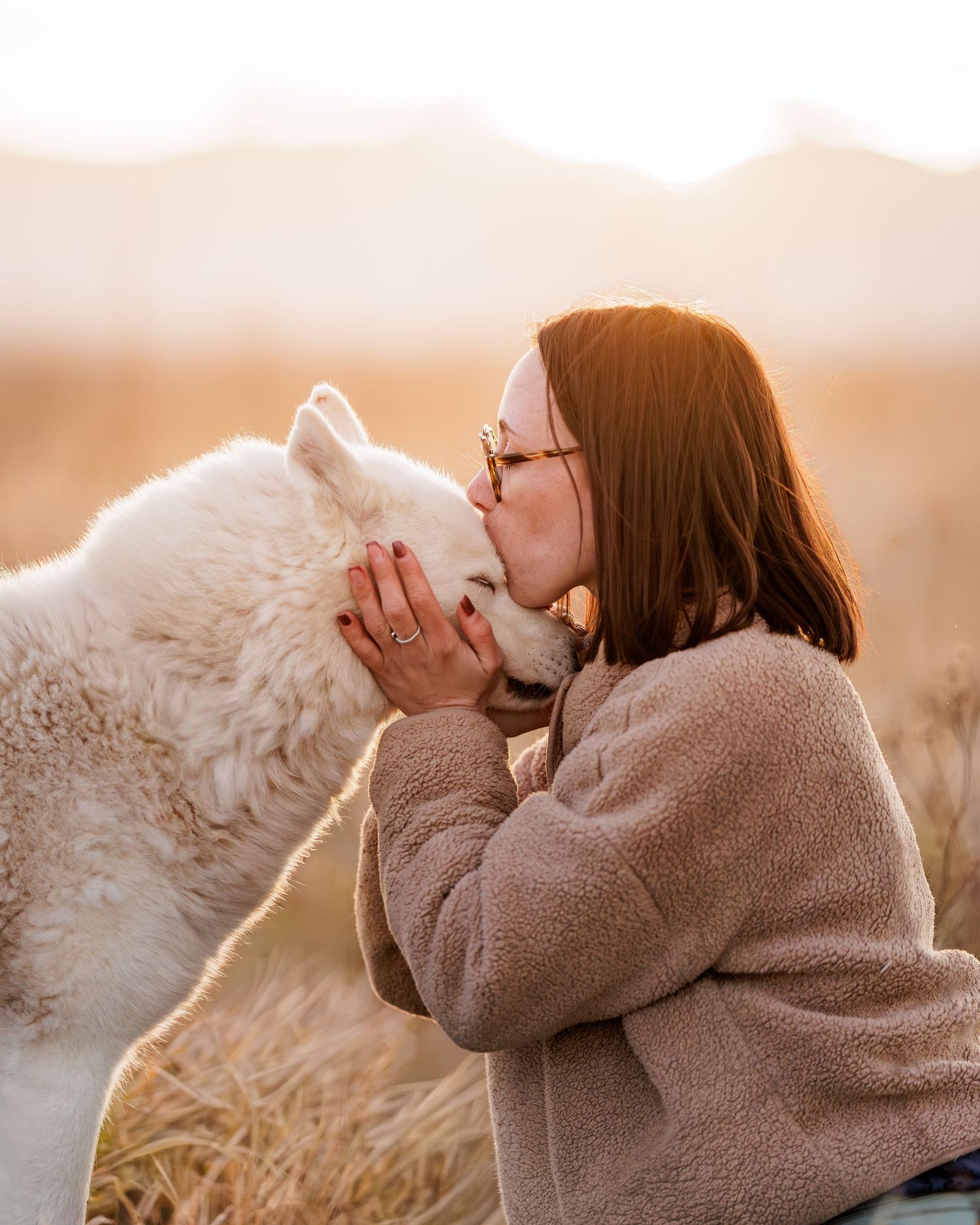 A woman with red hair kisses a fluffy white dog outdoors in golden sunset light.