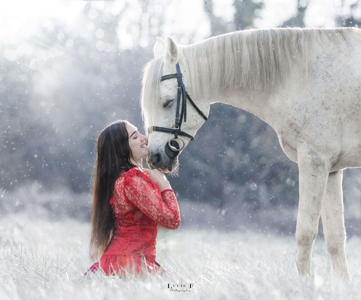 A person in a flowing red dress poses artistically with a white horse in a snowy winter landscape.