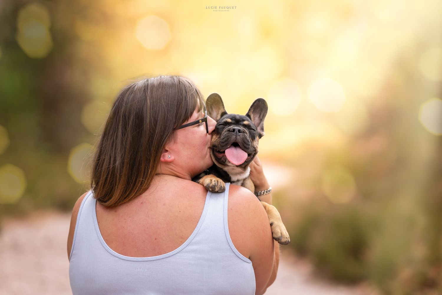 Woman in white tank top kissing a happy French Bulldog puppy outdoors in golden sunlight.