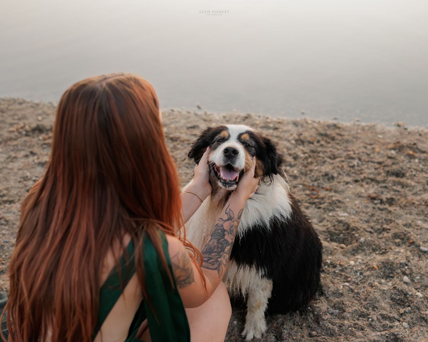 Red-haired tattooed woman petting a happy black and white Australian Shepherd dog outdoors near water.
