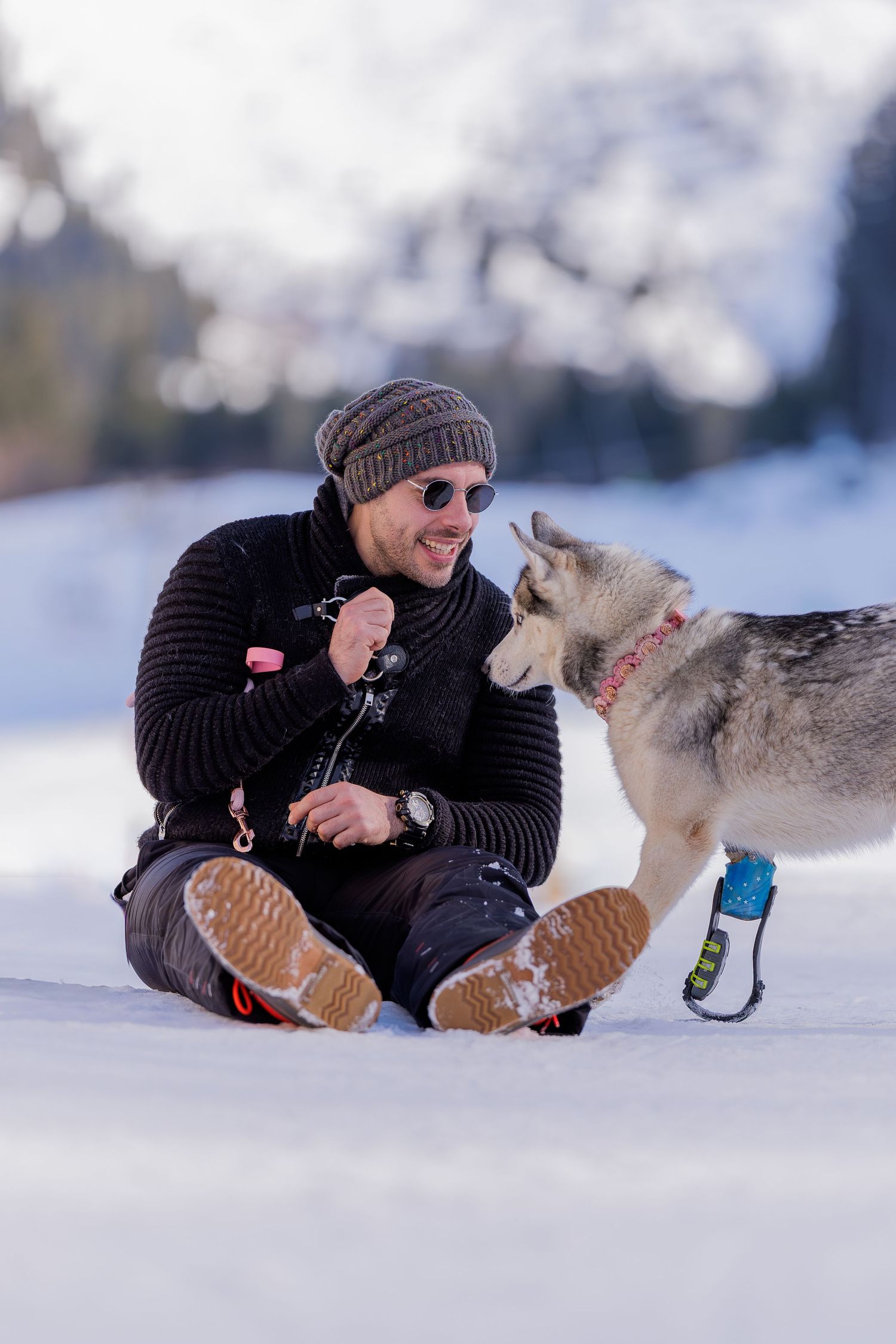 Person lying in snow wearing black sweater and beanie plays with friendly lynx cat in winter setting.
