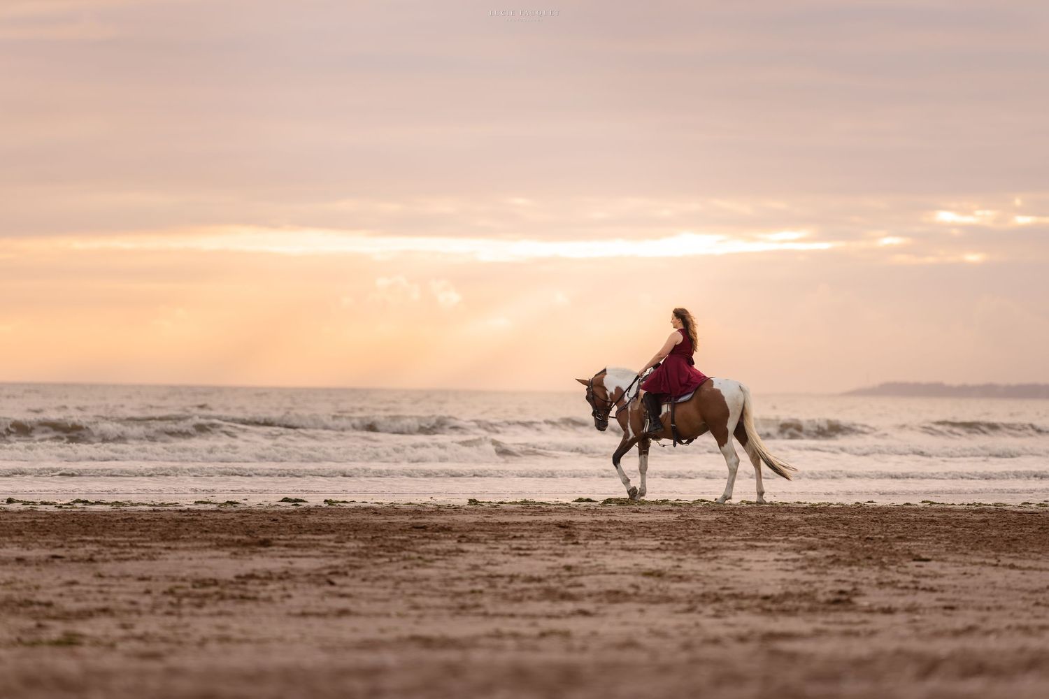 Woman in red dress riding a white horse on a sandy beach at sunset with ocean waves in the background.