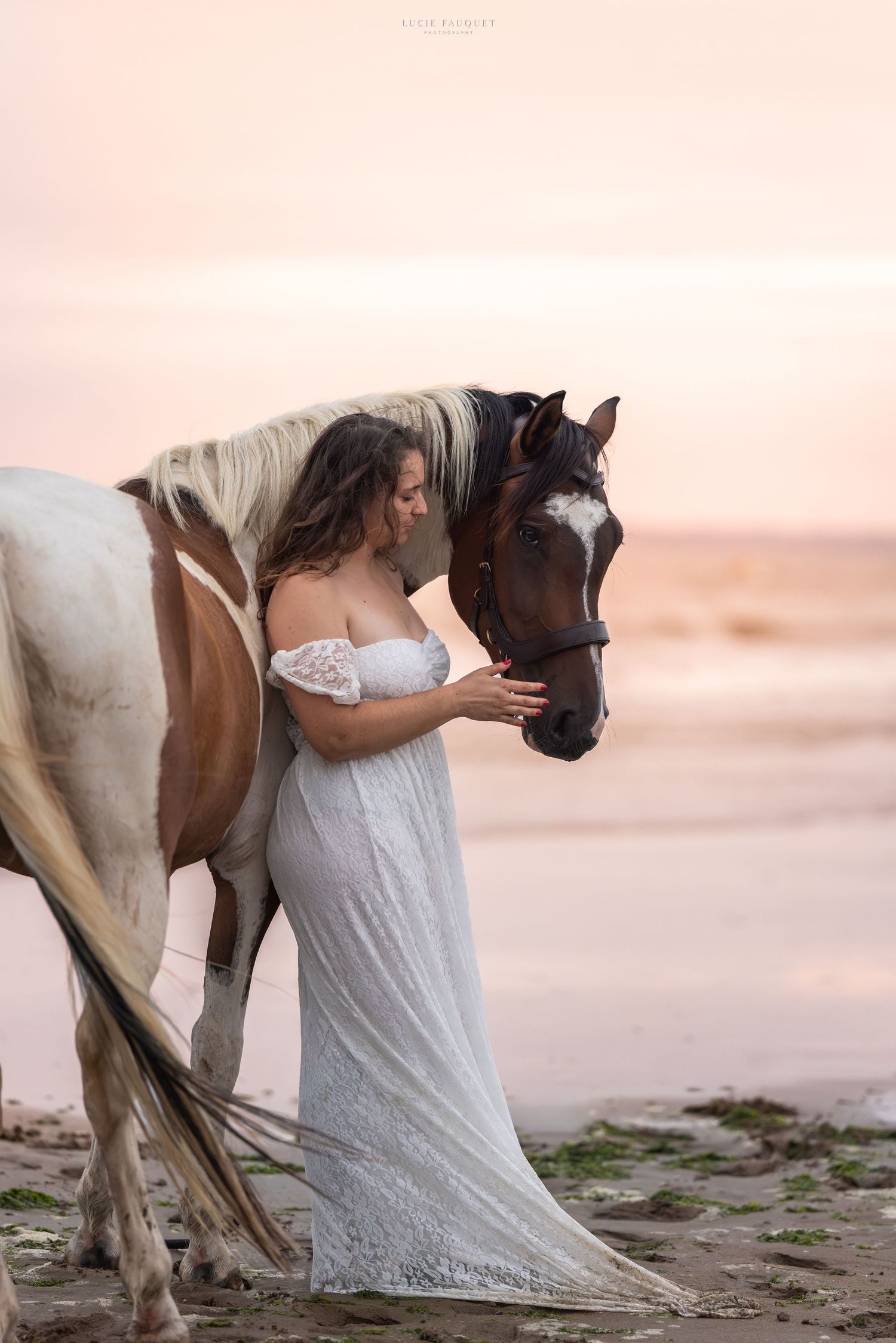 Woman in white lace gown tenderly embraces a paint horse on a sandy beach at sunset.