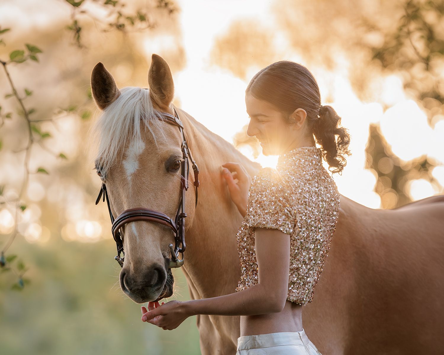Young woman in sequin top feeding a palomino horse at golden sunset in a serene outdoor setting.