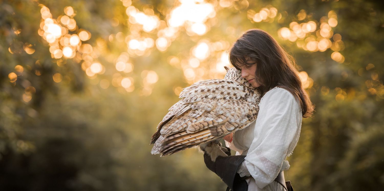 Young woman in white blouse holding a large owl in a sunlit forest with golden bokeh background.