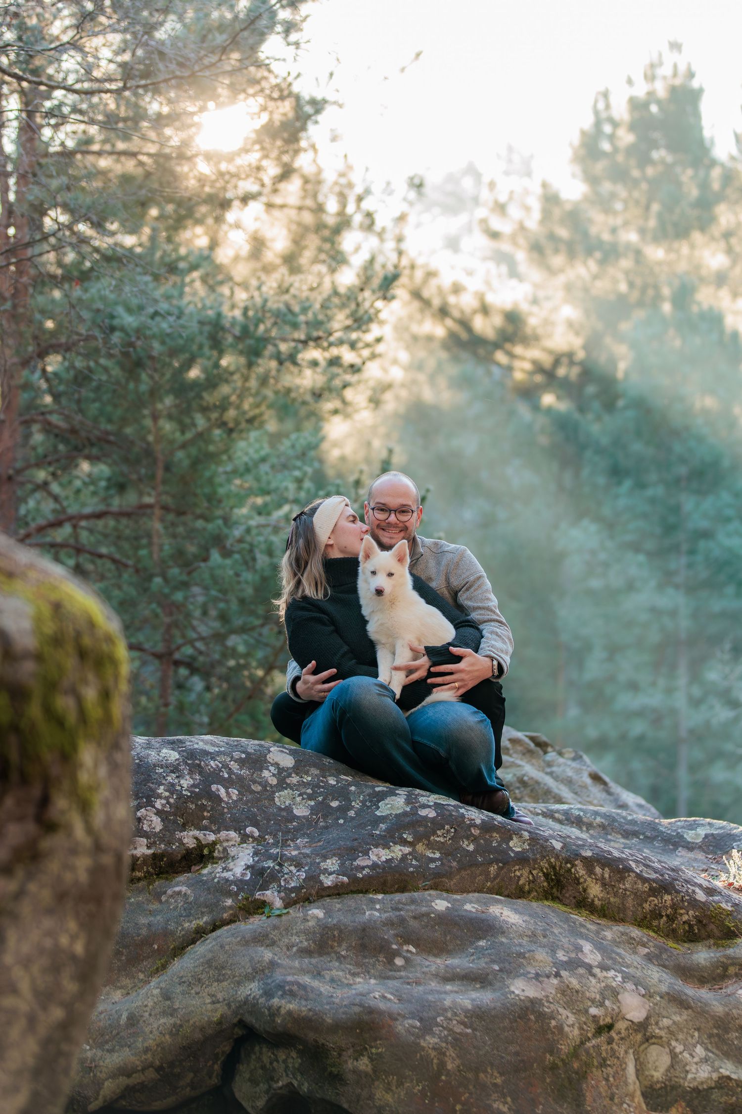 Couple sitting on mossy boulder in misty forest with small white dog as sunlight filters through pine trees.
