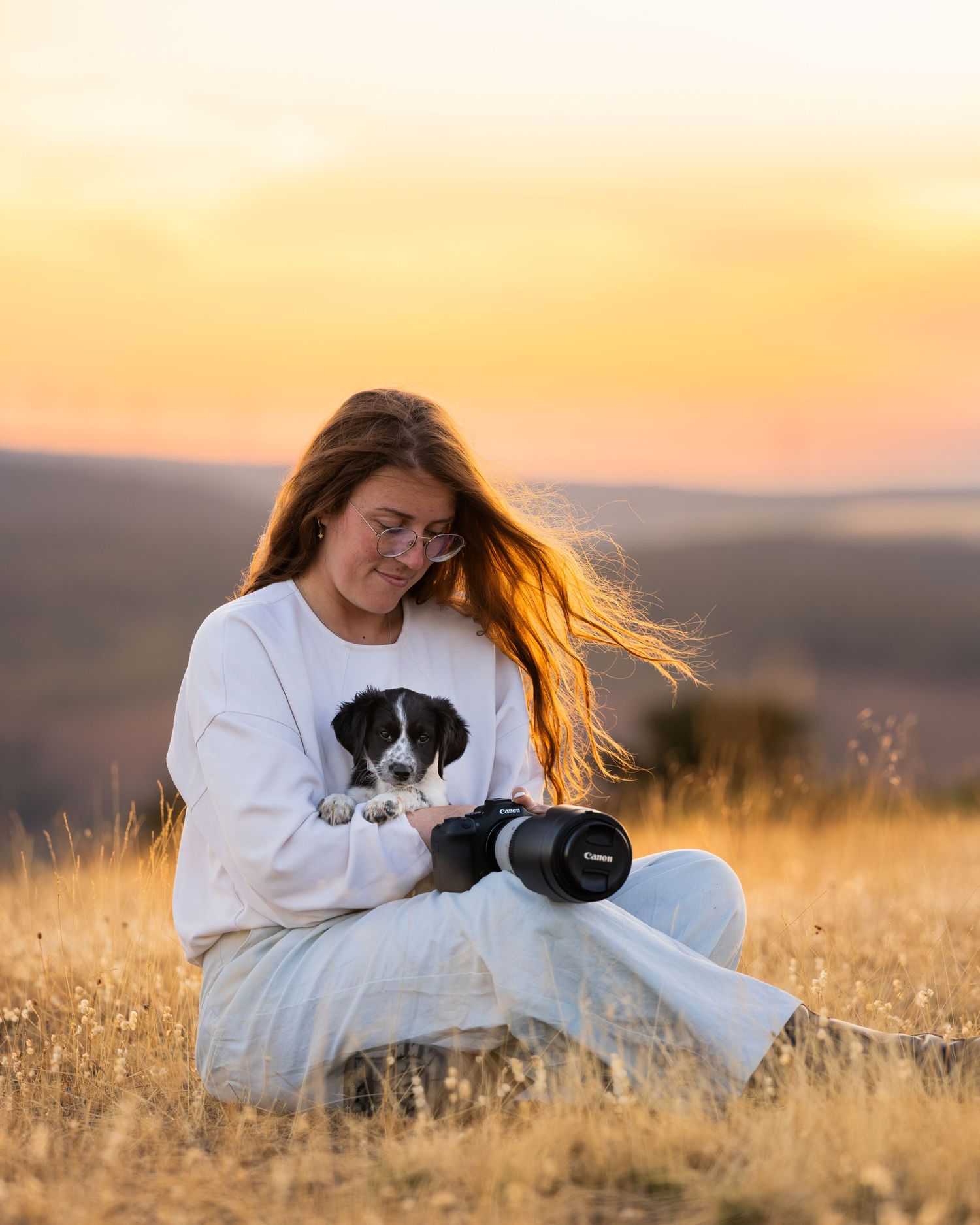 Woman sitting in golden field at sunset, holding a Canon camera, wearing white sweatshirt with puppy print, hair blowing in wind.