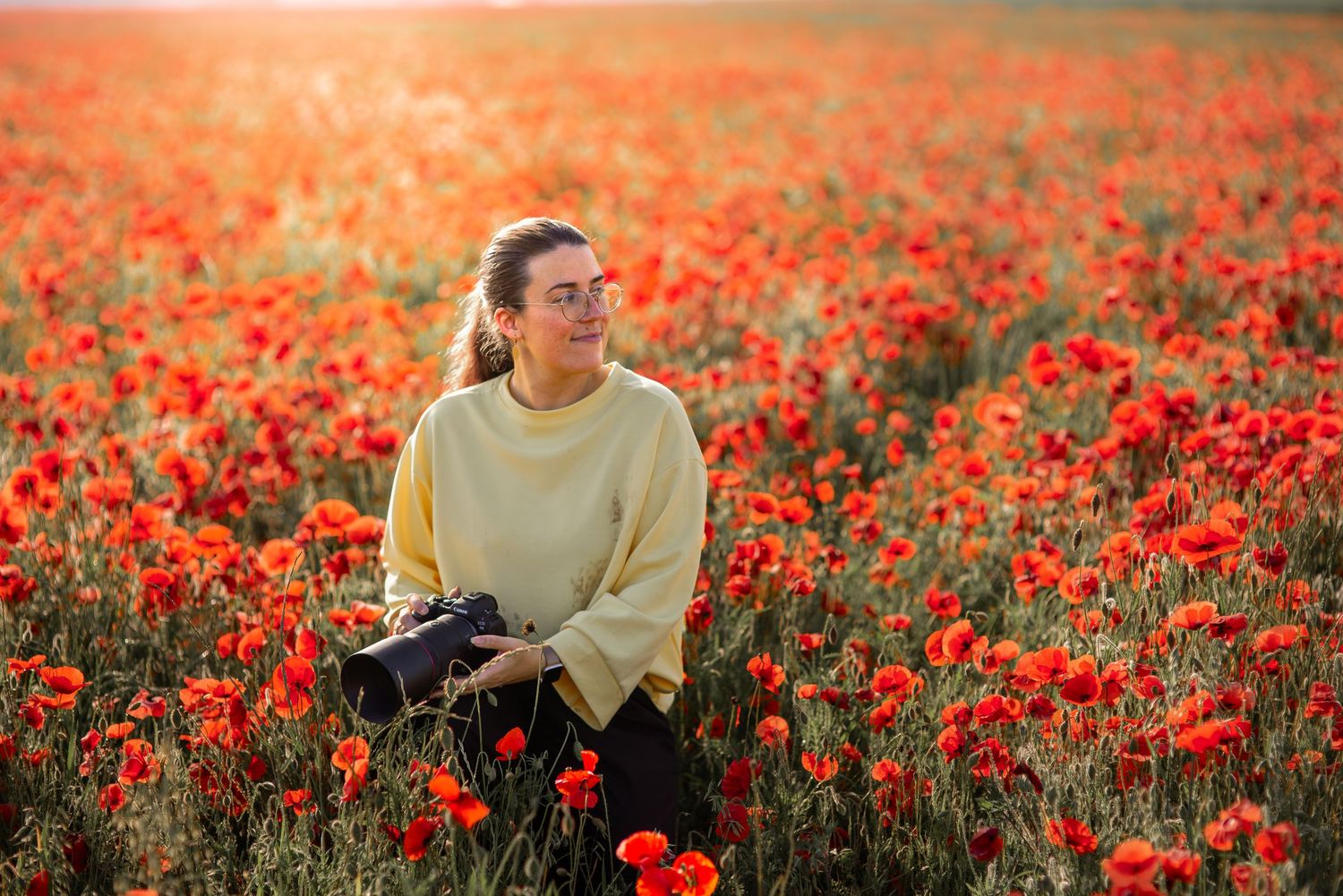 Young woman in yellow sweater holding camera in vibrant red poppy field during golden hour.