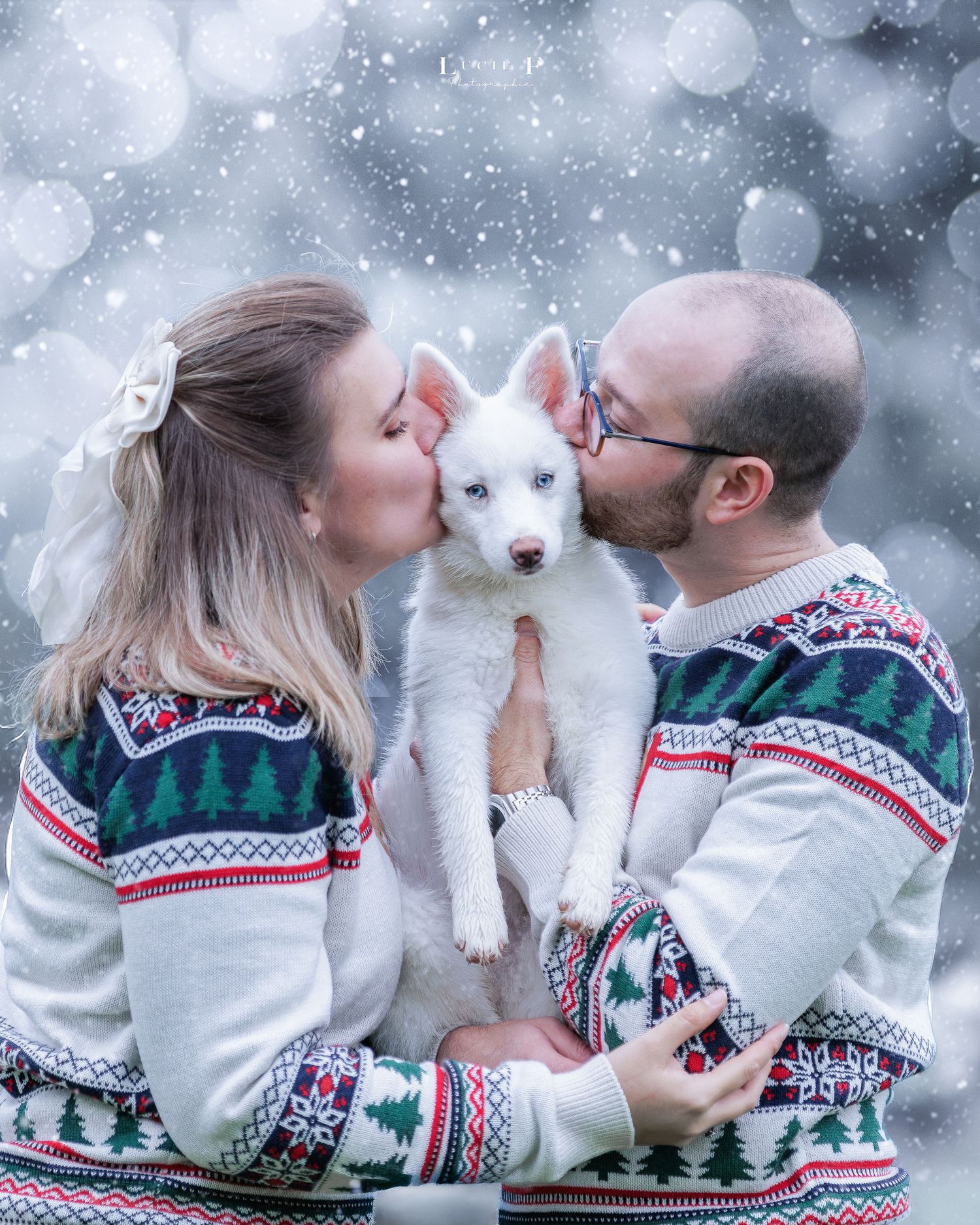 Couple in matching Christmas sweaters kissing a white husky puppy in a snowy bokeh background.