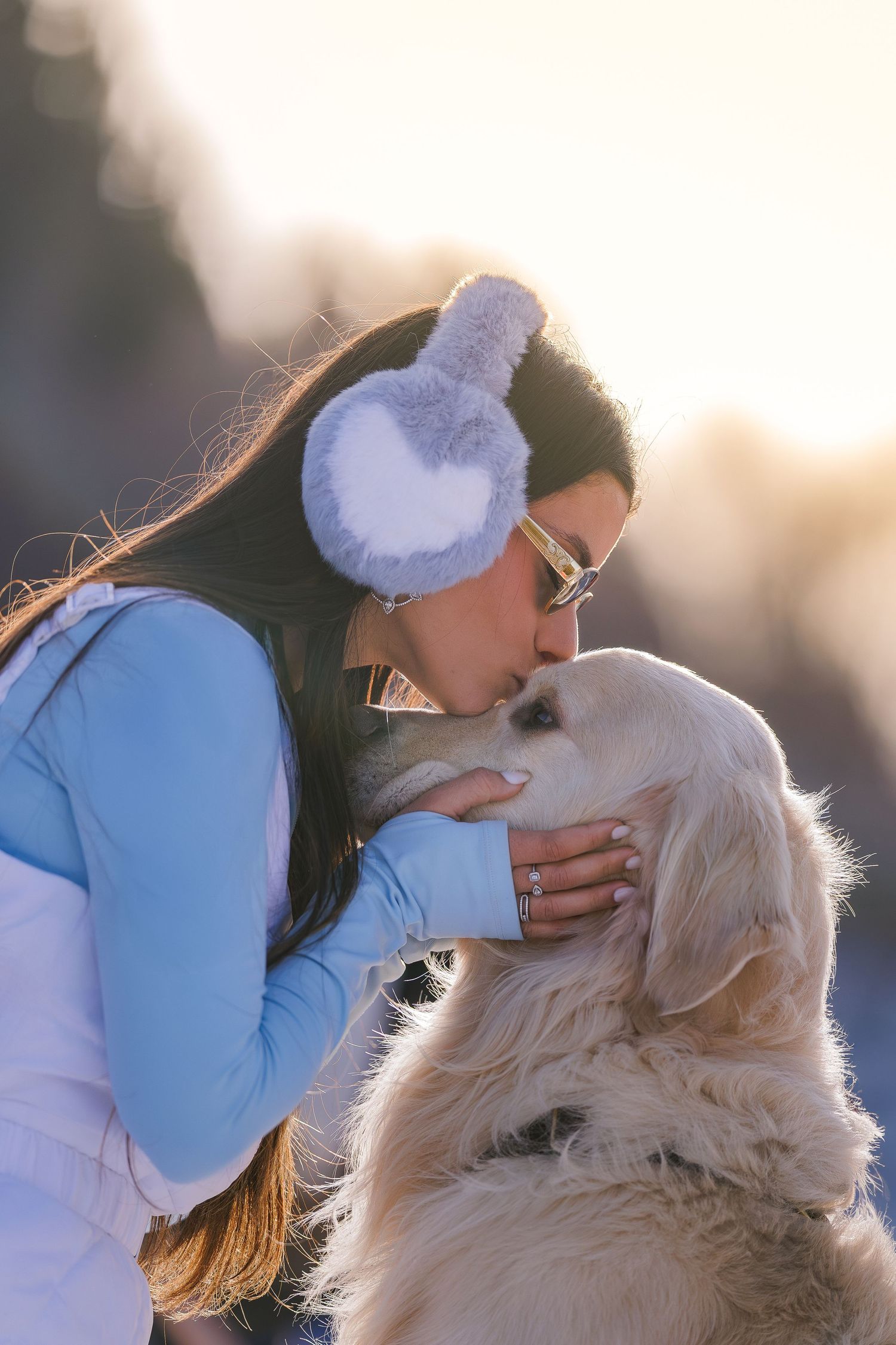 Woman in blue jacket and fuzzy earmuffs kissing her golden retriever dog outdoors in winter snow.