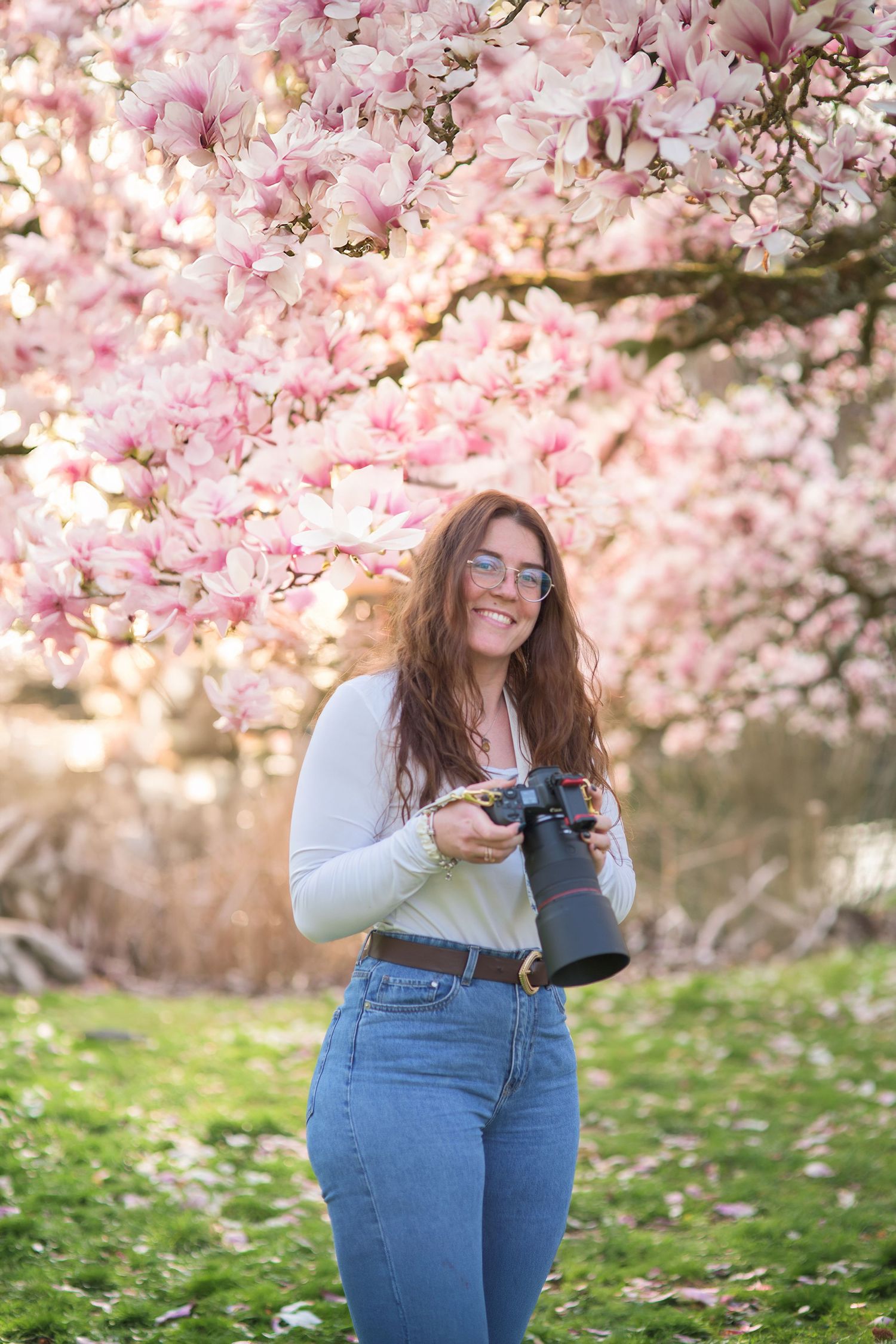 A person poses joyfully among blooming pink cherry blossom trees in springtime while wearing jeans and a light sweater.