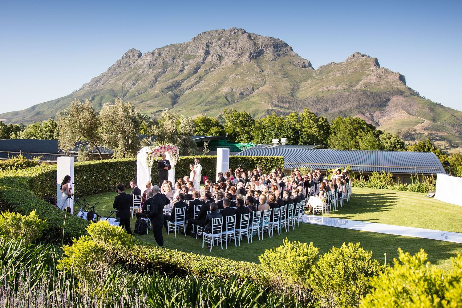 Outdoor destination wedding ceremony at Delaire Graff Estate, Stellenbosch, South Africa, with a dramatic mountain backdrop and blue sky.