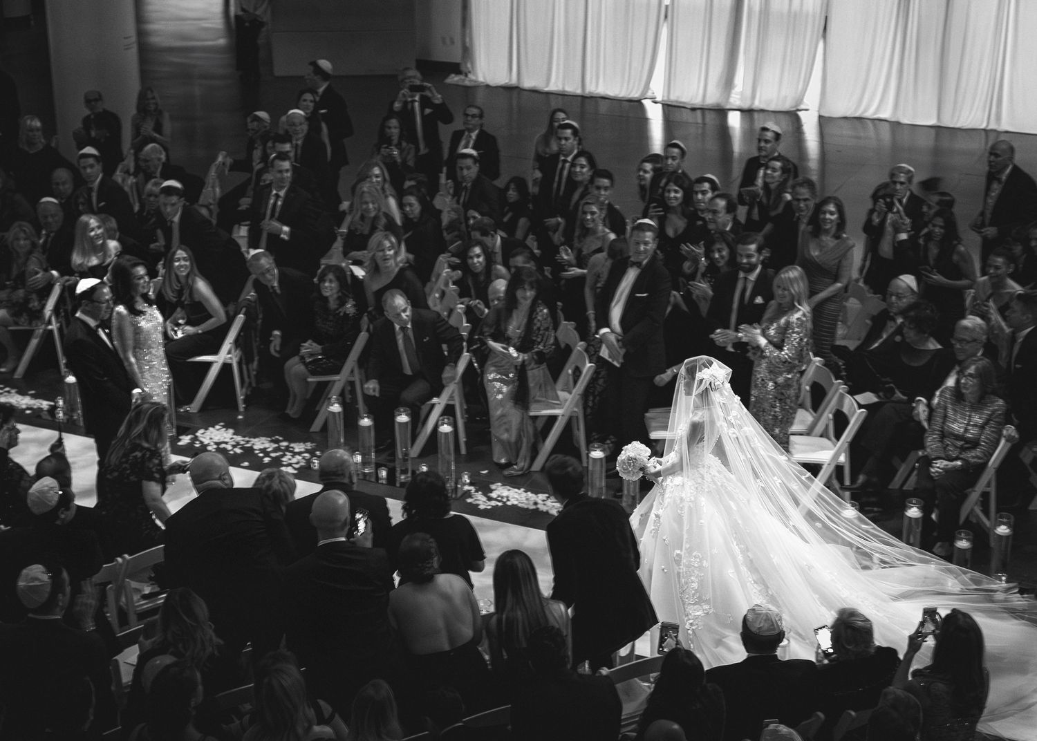 Black and white wedding ceremony photo showing bride in elegant gown walking down aisle with guests seated in the Brooklyn Museum of Art.
