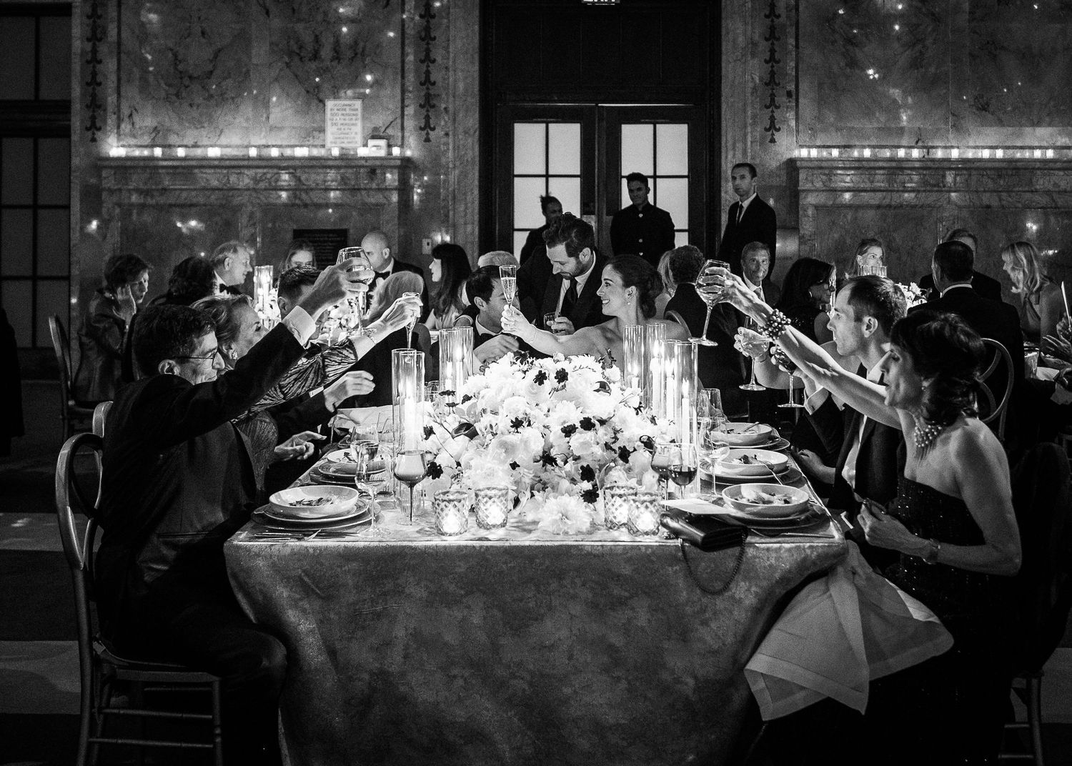 Black and white photo of a bride and groom toasting with guests at a candlelit wedding reception at the New York Public Library.