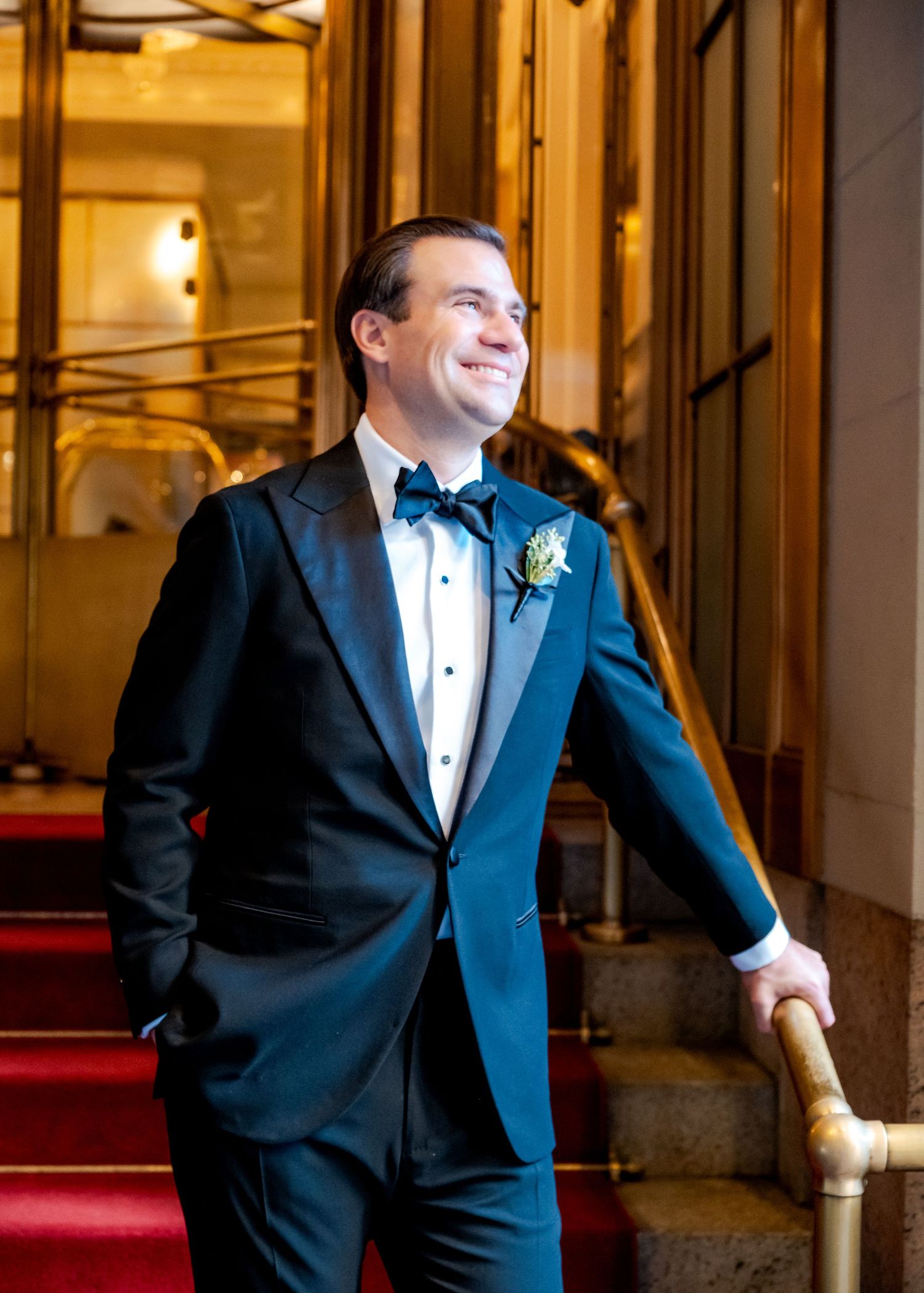 Smiling groom in navy tuxedo with bow tie and boutonniere poses on red carpeted staircase in elegant venue.
