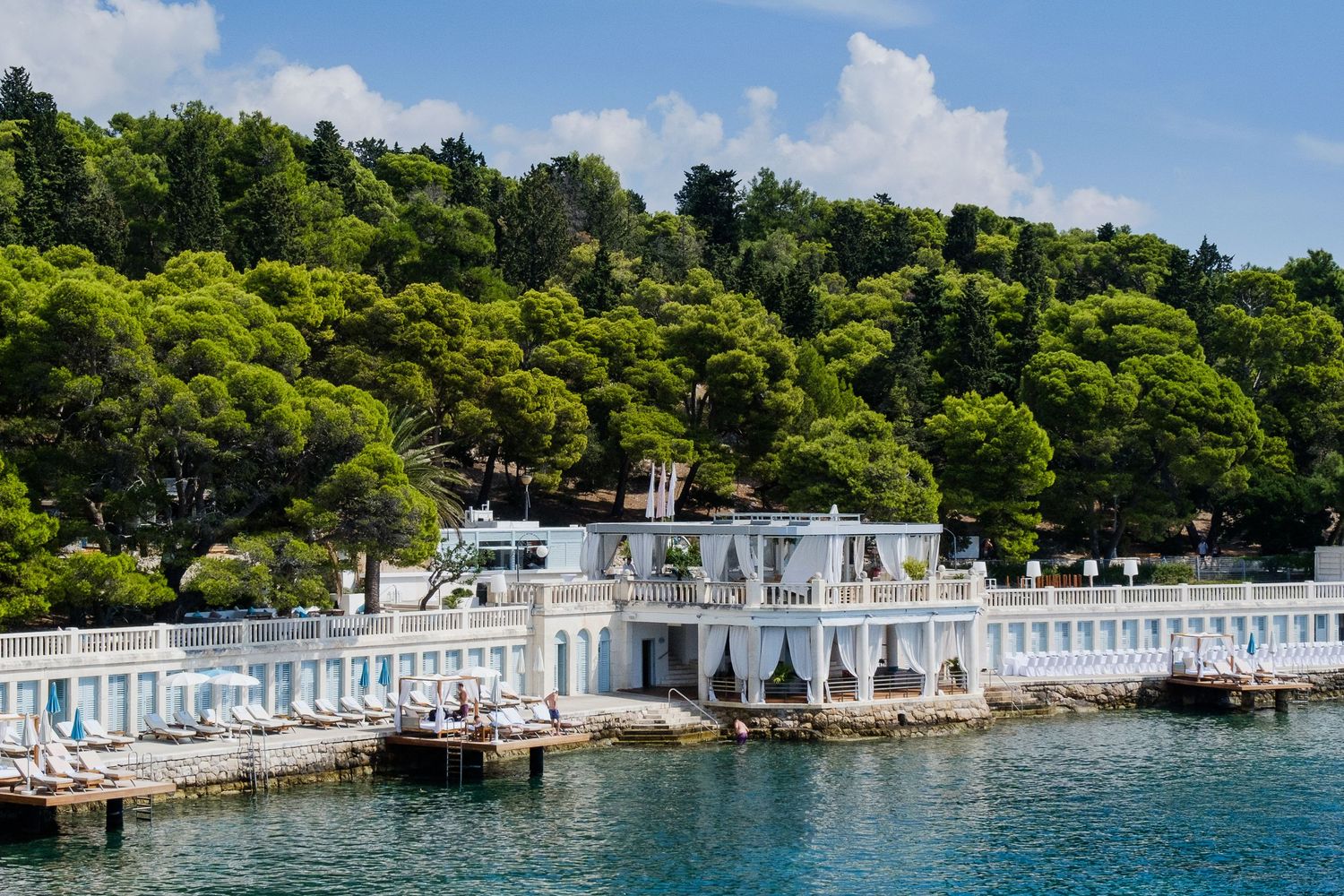 Elegant white waterfront building with arched colonnades nestled against lush green hillside trees by calm blue sea in Hvar, Croatia.