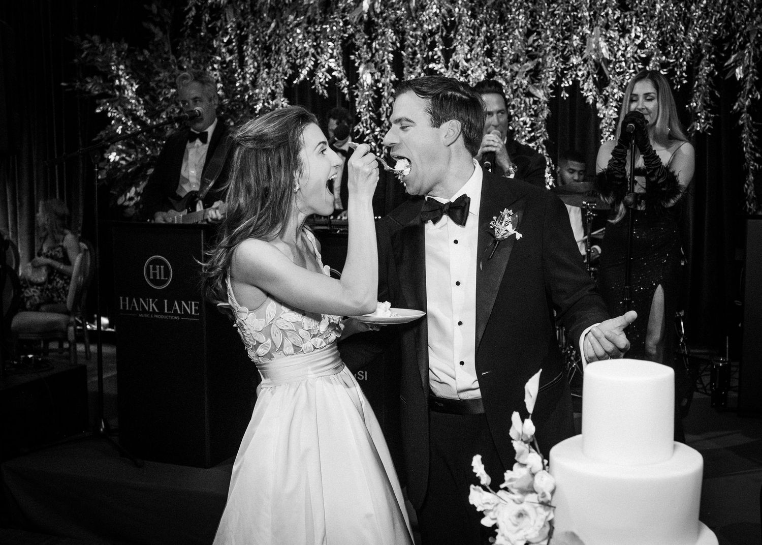 Bride feeds groom wedding cake at elegant black-tie reception with live band and floral backdrop.