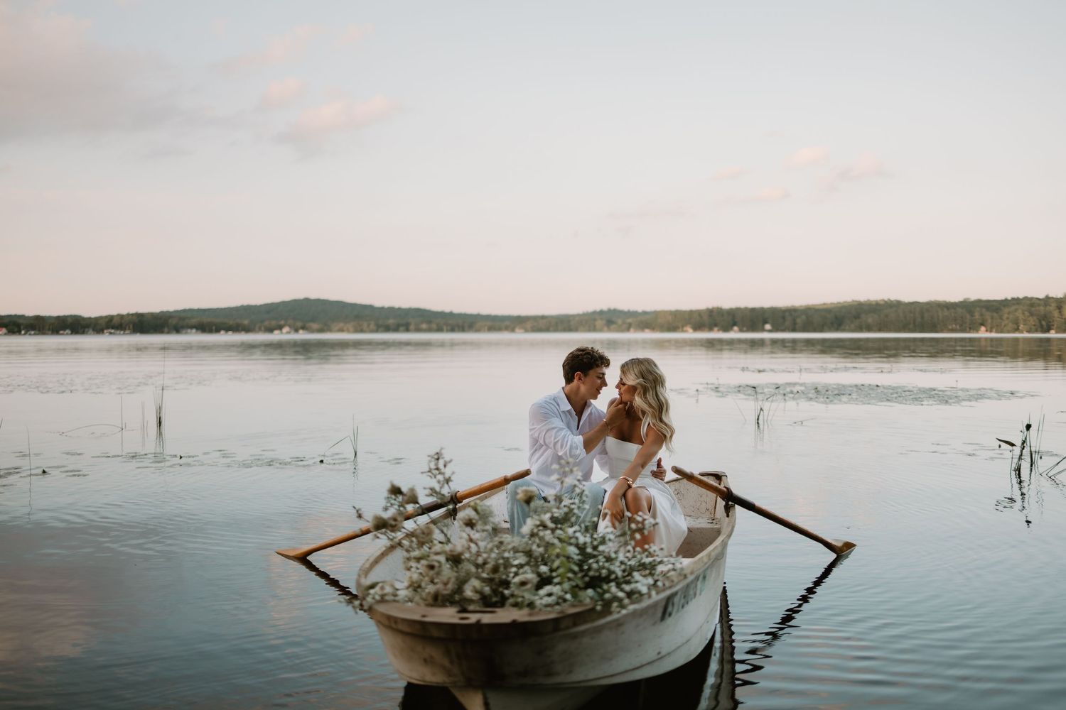 A romantic couple shares an intimate moment in a wooden rowboat decorated with flowers on a serene lake at dusk.