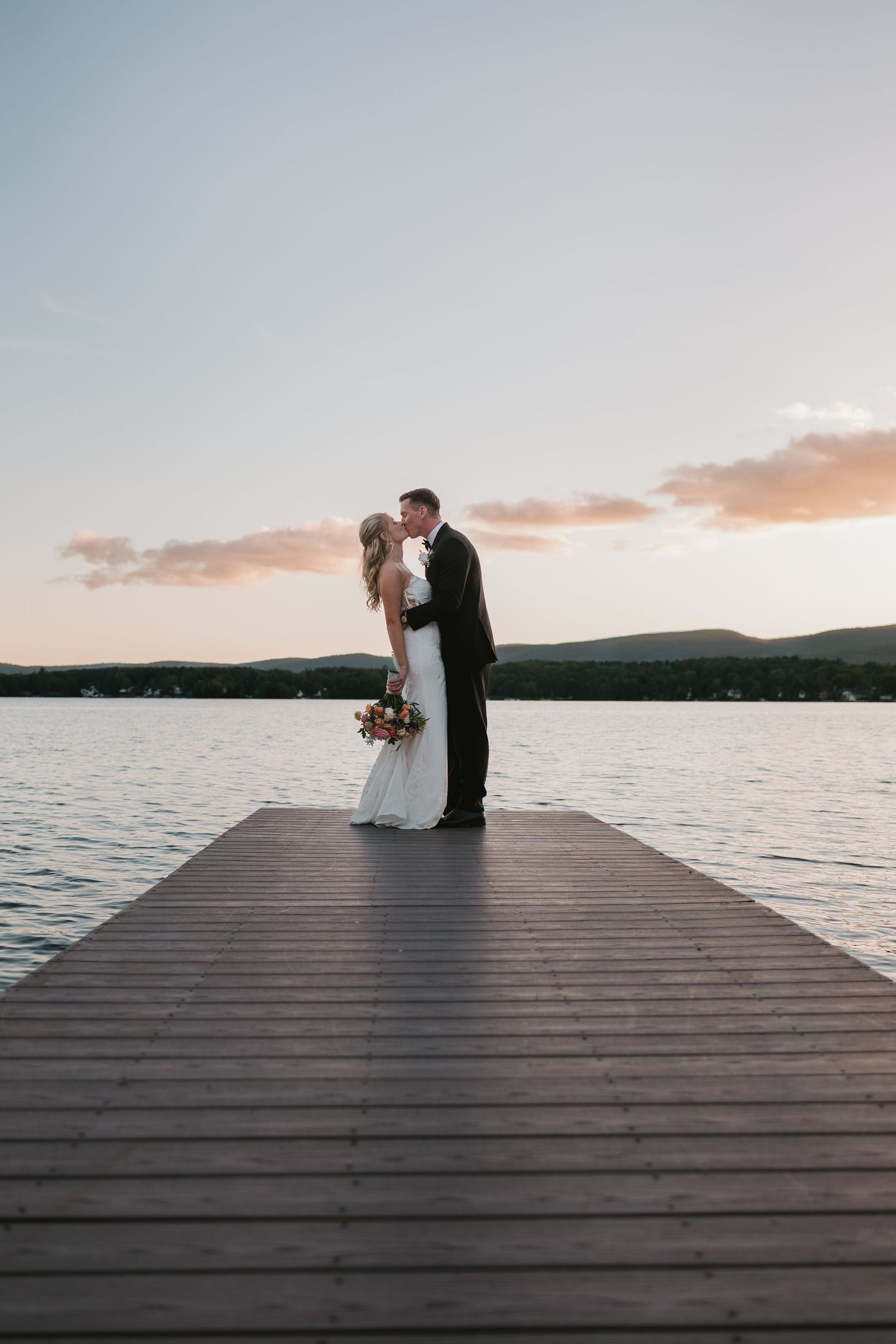 Bride and groom kissing on a wooden dock at sunset with a serene lake and mountain backdrop.