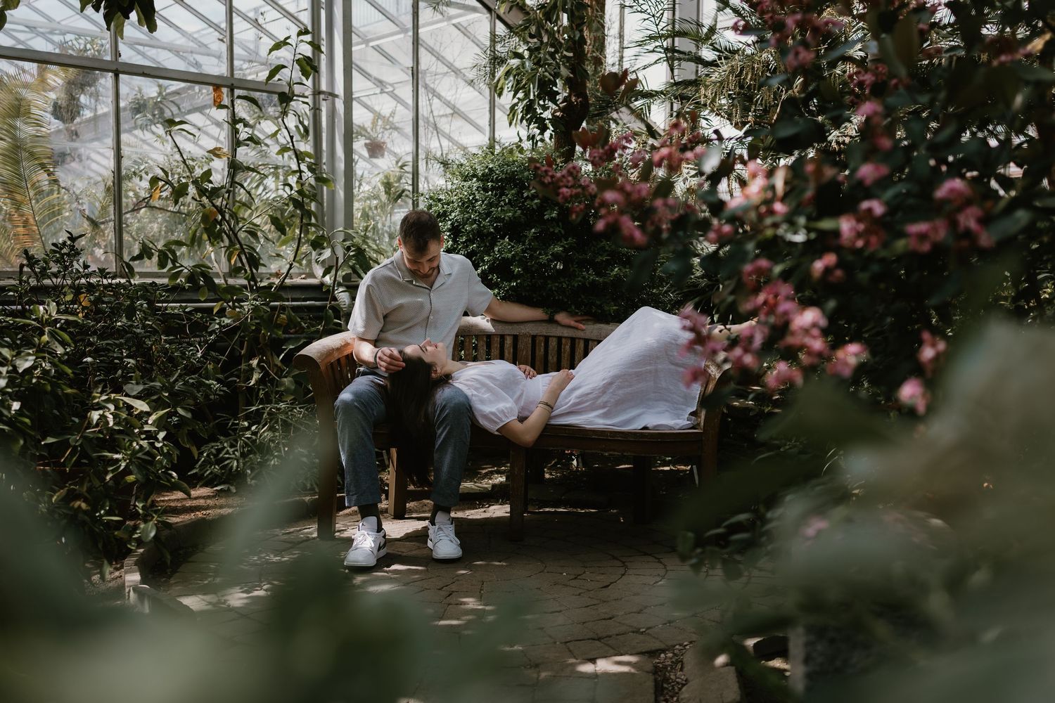 Couple sitting on a wooden bench surrounded by lush tropical plants and pink flowers inside a greenhouse.