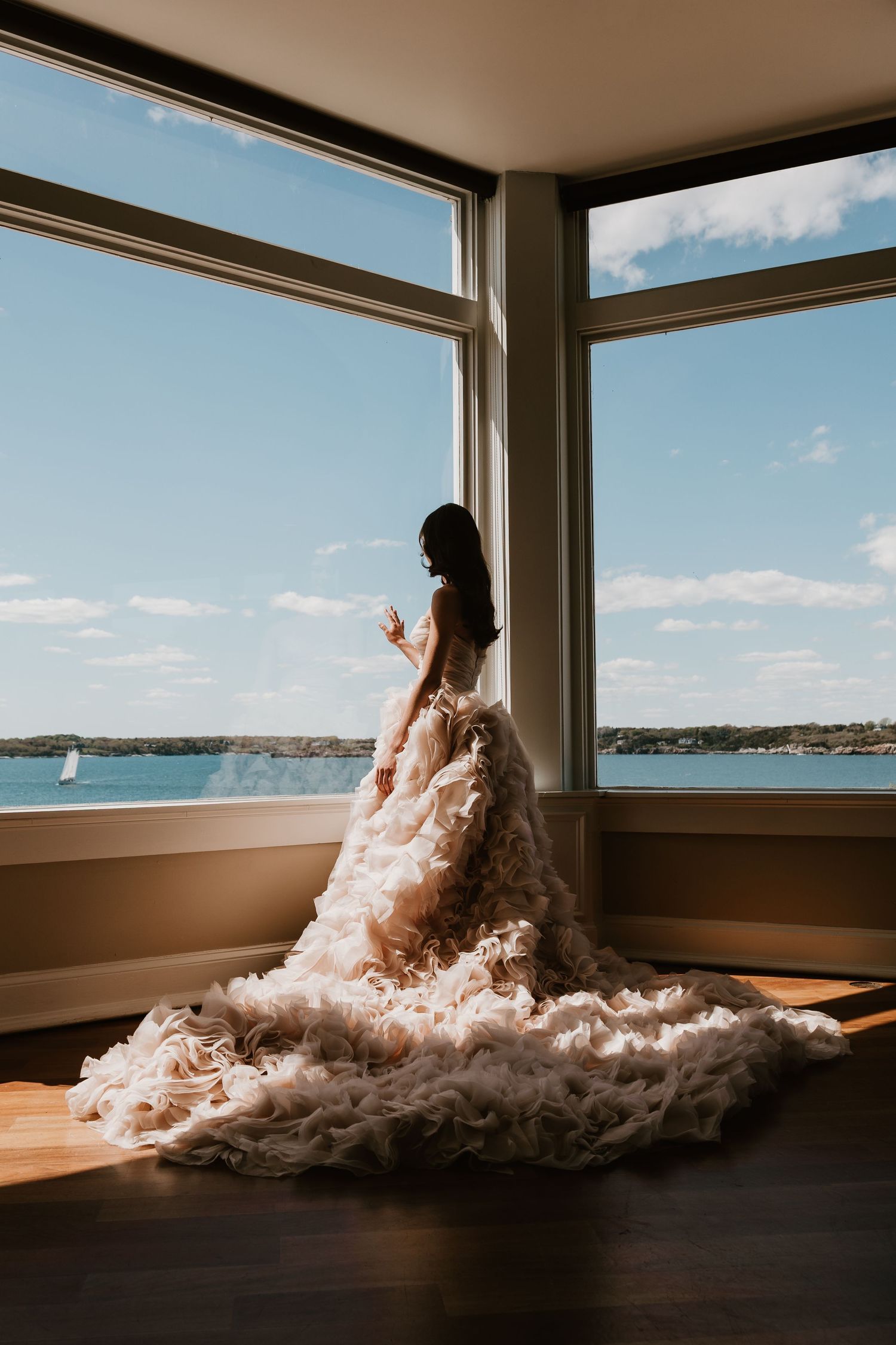 Bride in a ruffled ballgown stands by large corner windows overlooking a serene waterway on her wedding day.