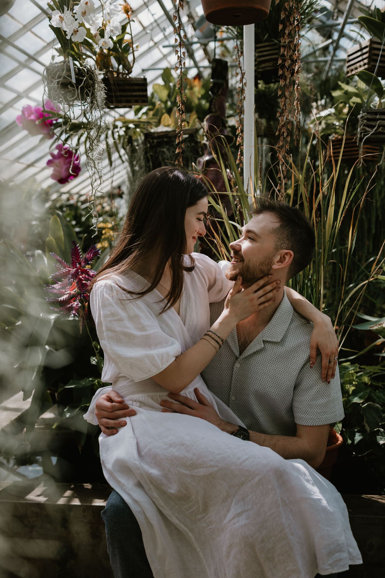 Couple sharing a romantic moment in a lush greenhouse, woman in white dress touching man's face.