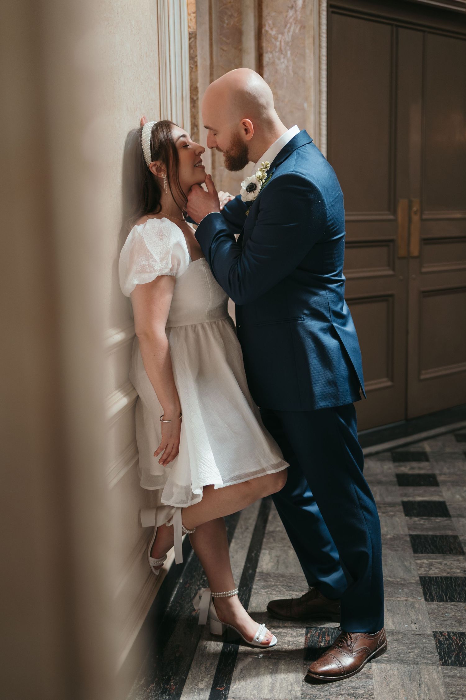 Bride in white mini dress and groom in blue suit share a romantic moment leaning against a wall.