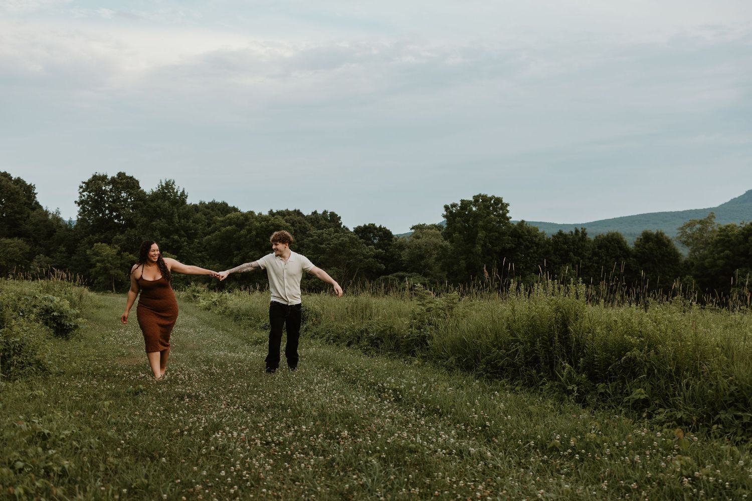 A couple runs hand-in-hand through a lush green meadow with trees and mountains in the background.