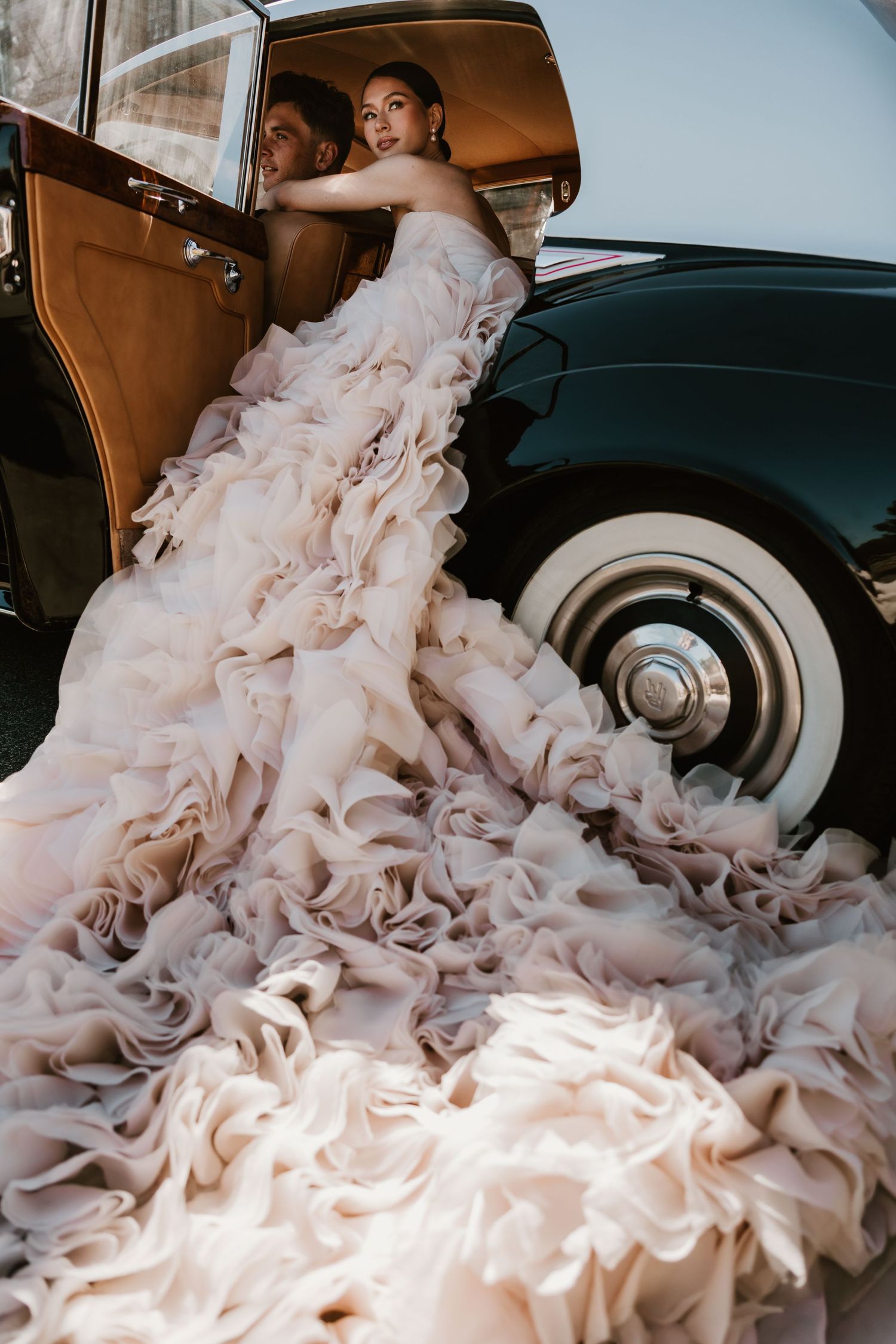 Bride in a voluminous blush ruffled gown cascading from a vintage black car with white wall tires.