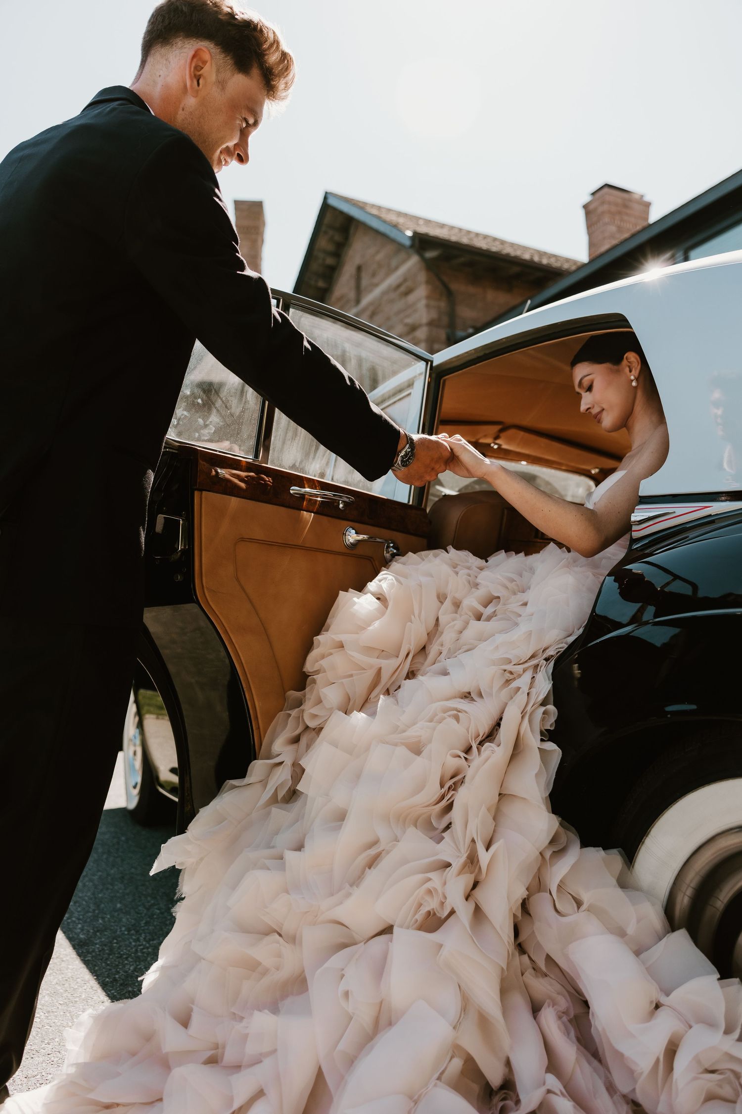 Groom in black suit helps bride in ruffled blush gown exit a vintage black car on their wedding day.