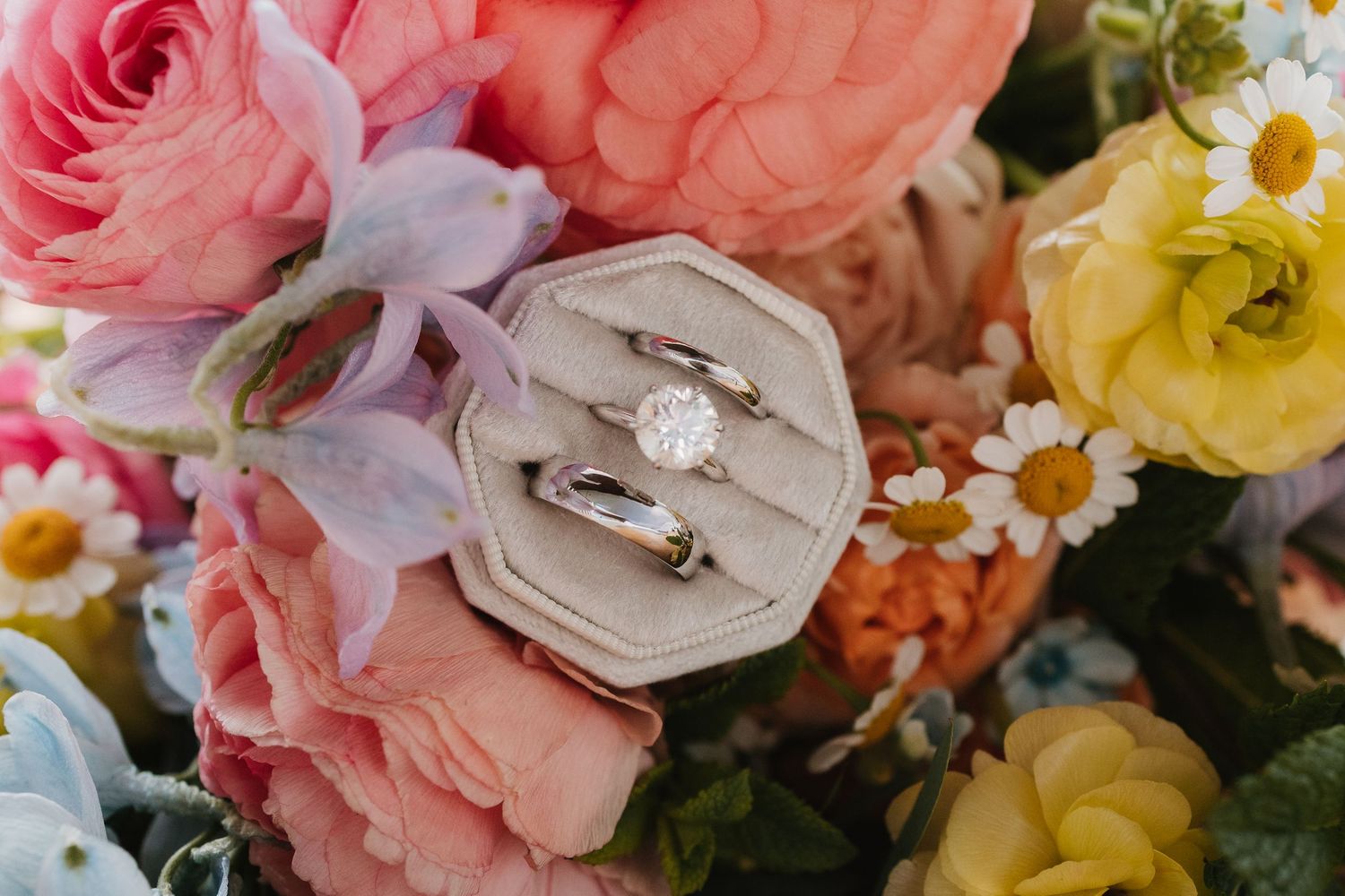 Wedding rings displayed in a velvet box surrounded by colorful spring flowers including pink roses and yellow blooms.