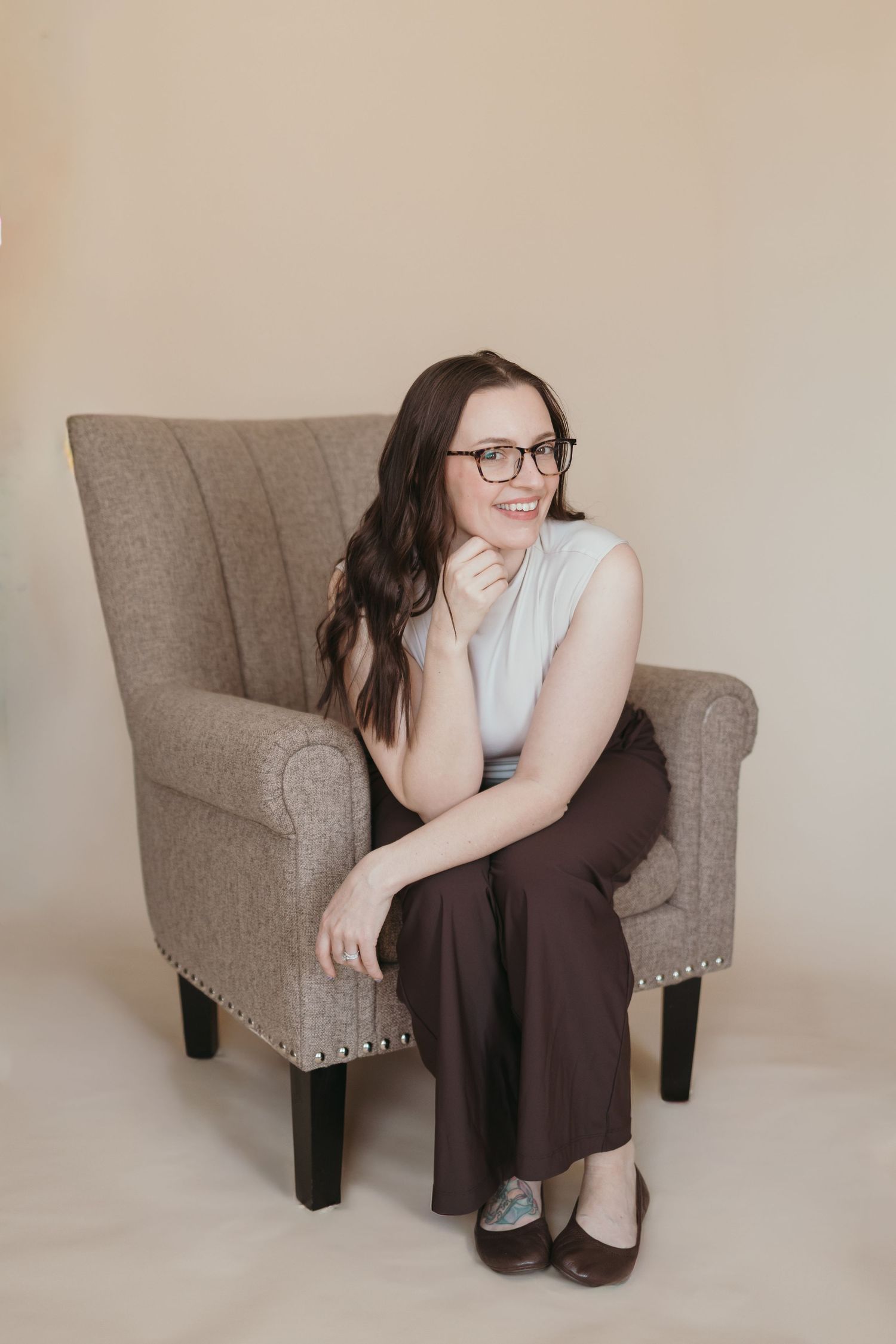 Woman with glasses sitting in gray armchair, wearing white top and brown pants, smiling in a neutral studio setting.