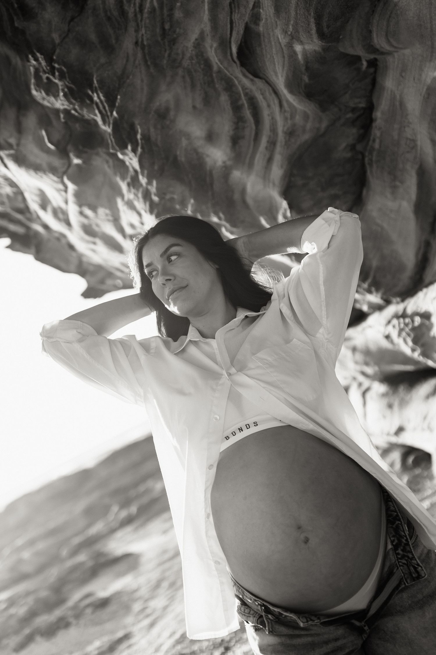 Black and white photo of pregnant woman in open white shirt exposing baby bump, posing against rocky background.