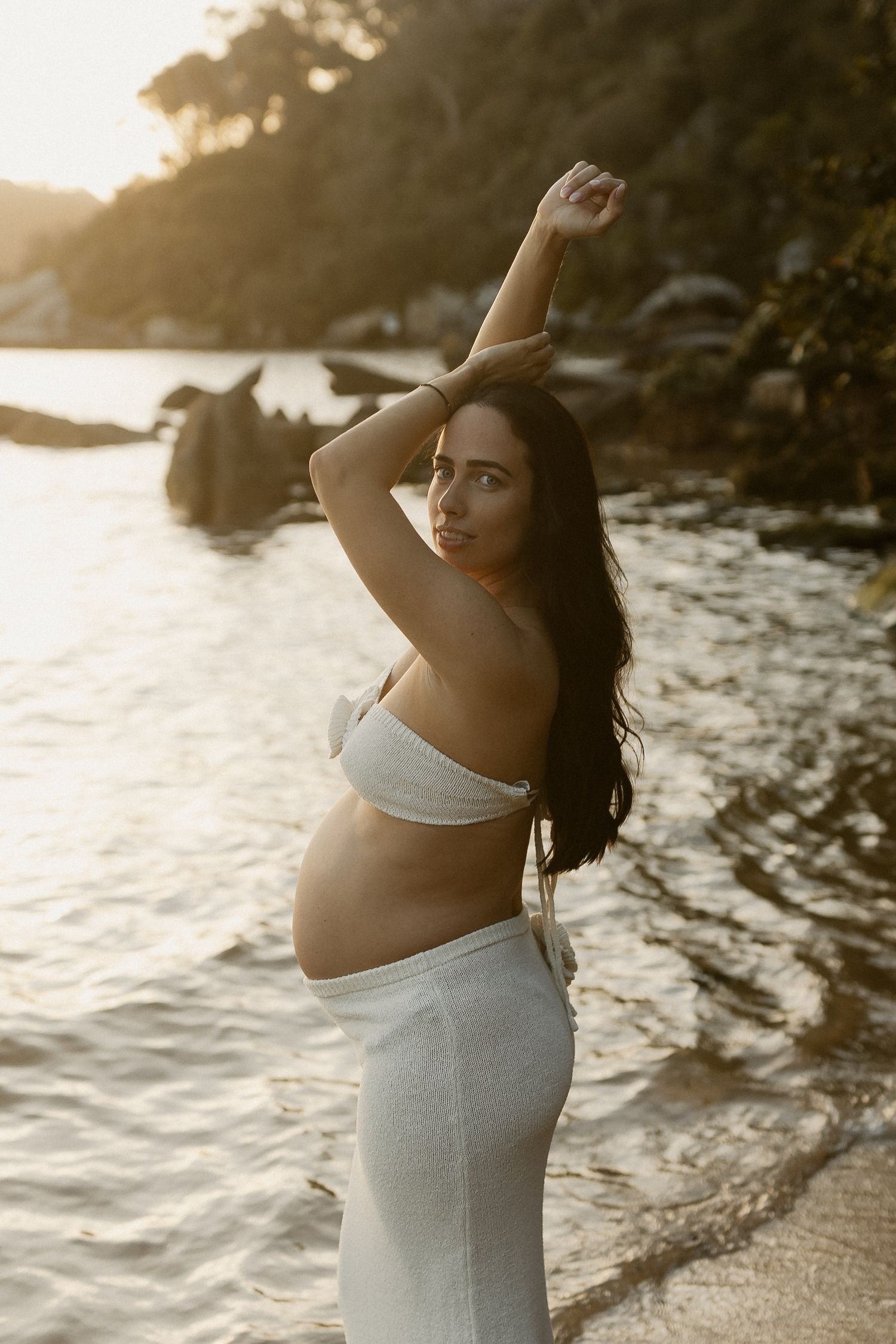 Expectant mother in white attire poses gracefully by the ocean during sunset.