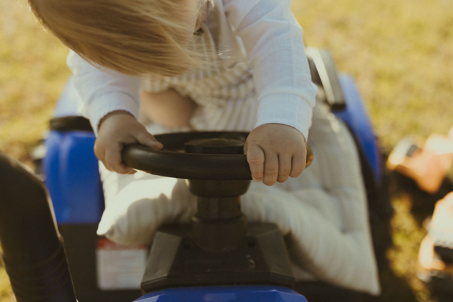 Small hands grip the steering wheel of a blue ride-on toy vehicle in warm sunlight.