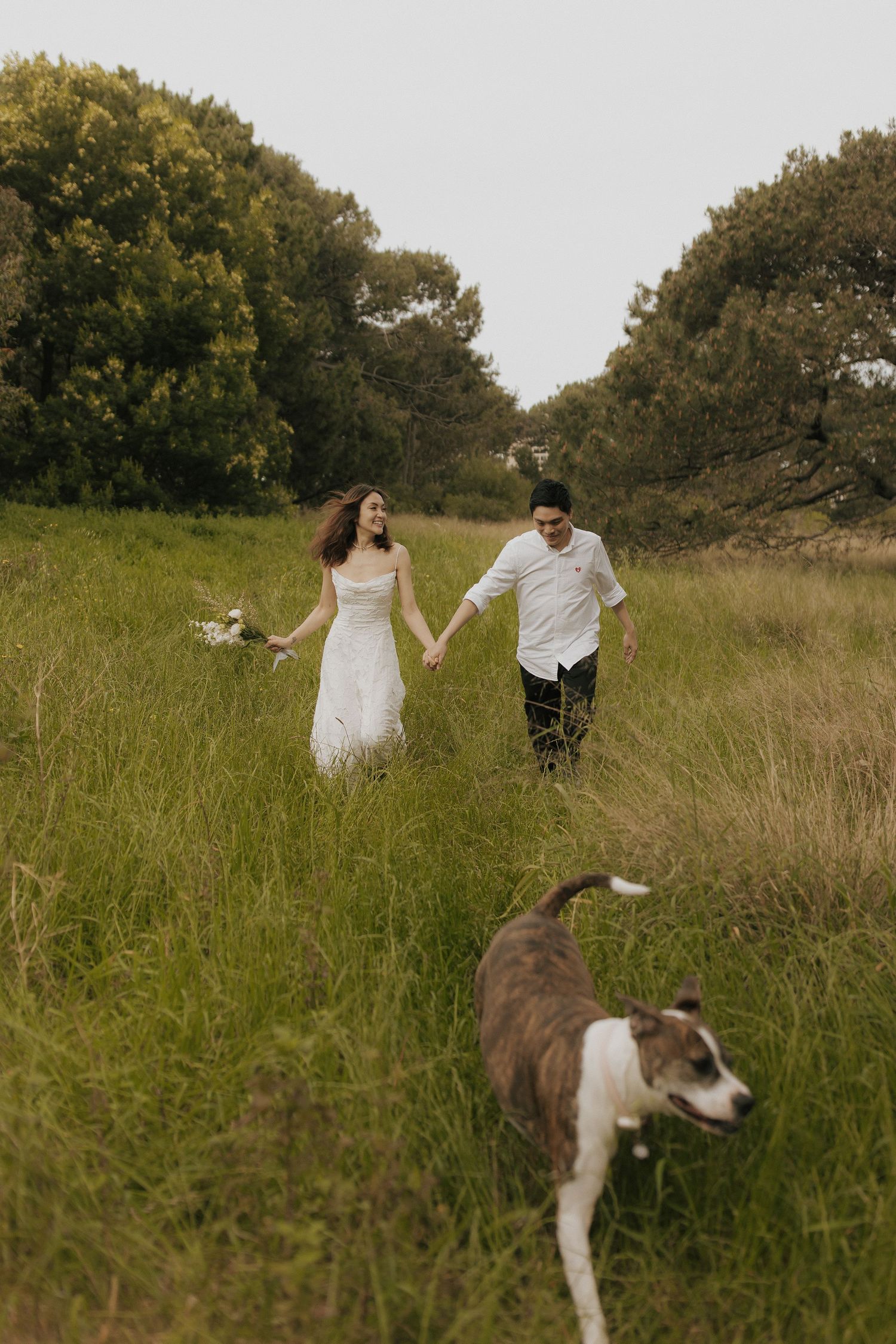 A couple walks through tall grass in a meadow while being followed by their dog.