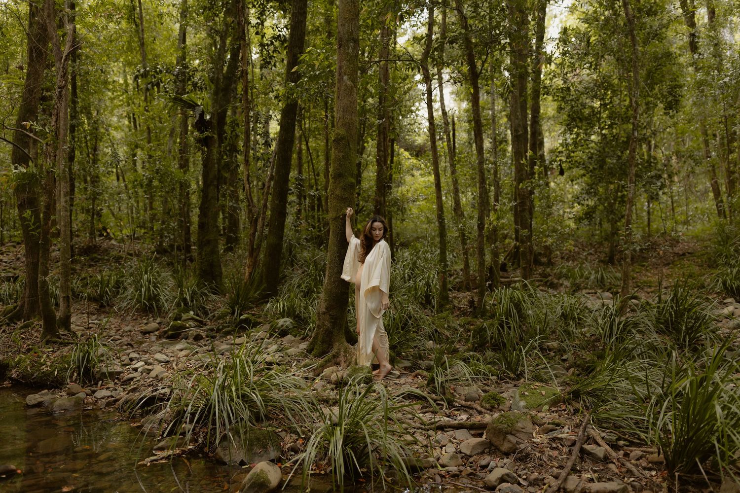 A couple shares an intimate moment while standing along a creek in a lush green forest setting.